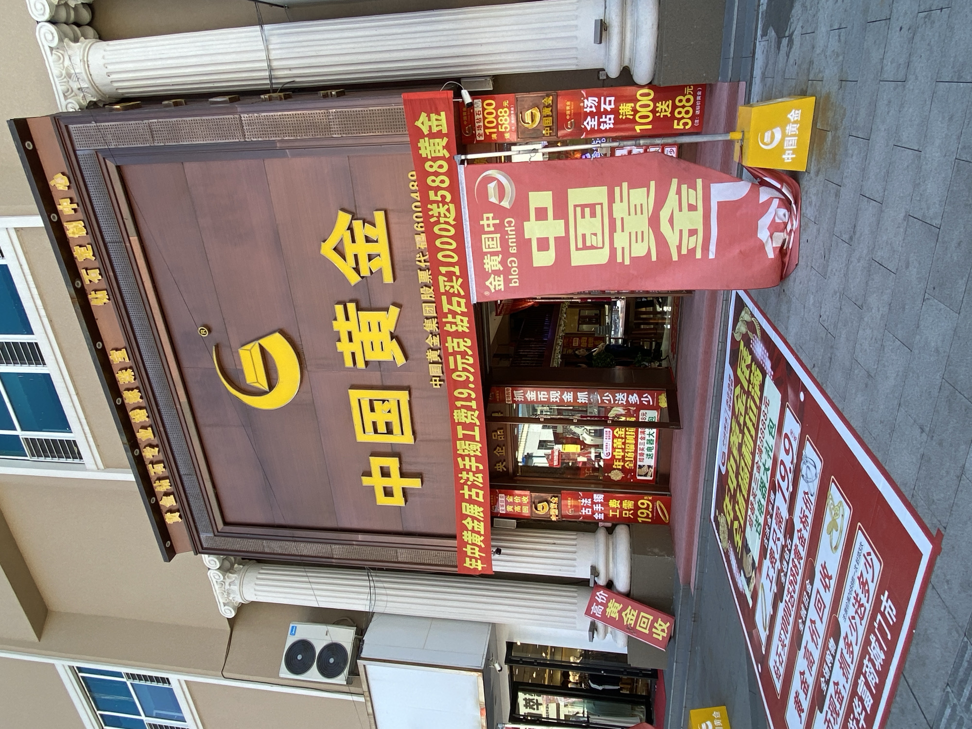 The image shows the exterior of a "China Gold" jewelry store located on Xing'an Street, China. The store has a prominent brown facade with large yellow Chinese characters spelling out "中国黄金" (China Gold) above the entrance. A red banner hangs across the storefront with promotional text in Chinese. Several smaller red and yellow signs are displayed, some on the ground and others near the entrance, advertising sales and services. One large red banner in front of the store also features Chinese text and a logo. White columns flank the entrance, and an air conditioning unit is visible on the building's exterior. The store appears to be open, with the entrance doors visible, and a few people can be seen inside. The street in front of the store is paved with grey tiles, and some promotional materials are spread out on the ground.