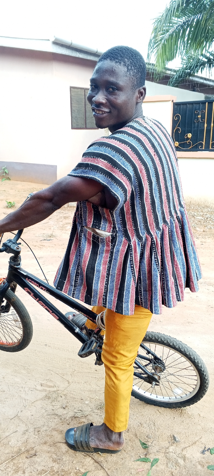 An adult male stands outdoors in Dzodze, Ghana, looking over his right shoulder at the viewer with a slight smile. He is dark-skinned with short dark hair, wearing a traditional Ghanaian smock featuring vertical black, red, and light blue stripes, yellow trousers, and dark slide sandals. He holds the handlebars of a black BMX-style bicycle, possibly a Kent Chaos model, with his left hand. A coiled silver-colored U-lock is attached to the bicycle's frame. The man's left foot is on the dry, light brown ground, which exhibits sparse vegetation. In the background, a light-colored building with a brown-framed window and a white rain gutter is visible, along with a dark, ornate metal gate and green palm fronds against a bright sky.