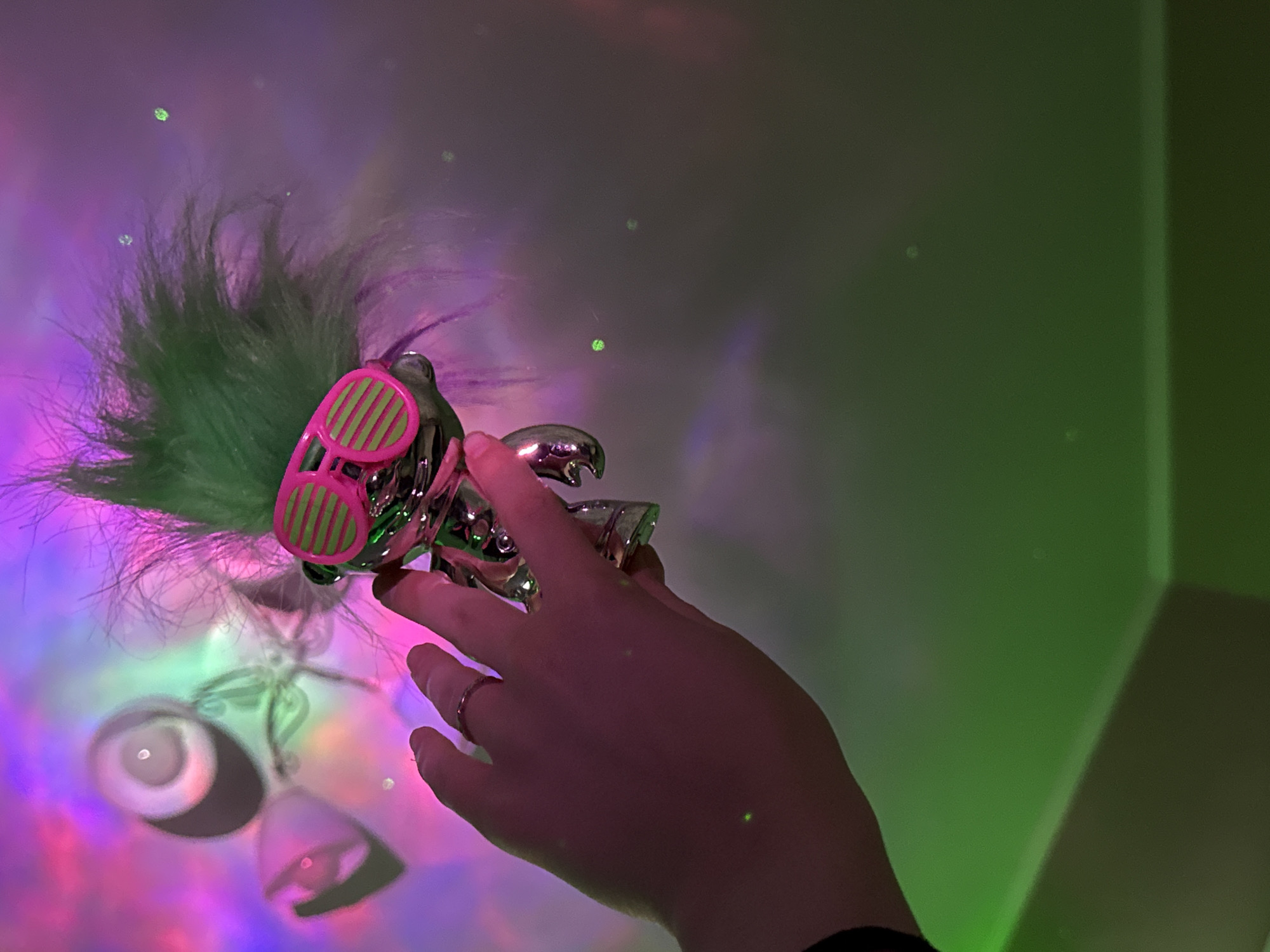 A hand is holding a silver troll doll with green hair and pink sunglasses against a wall illuminated with a mix of green, purple, and pink lights. The location is unspecified.