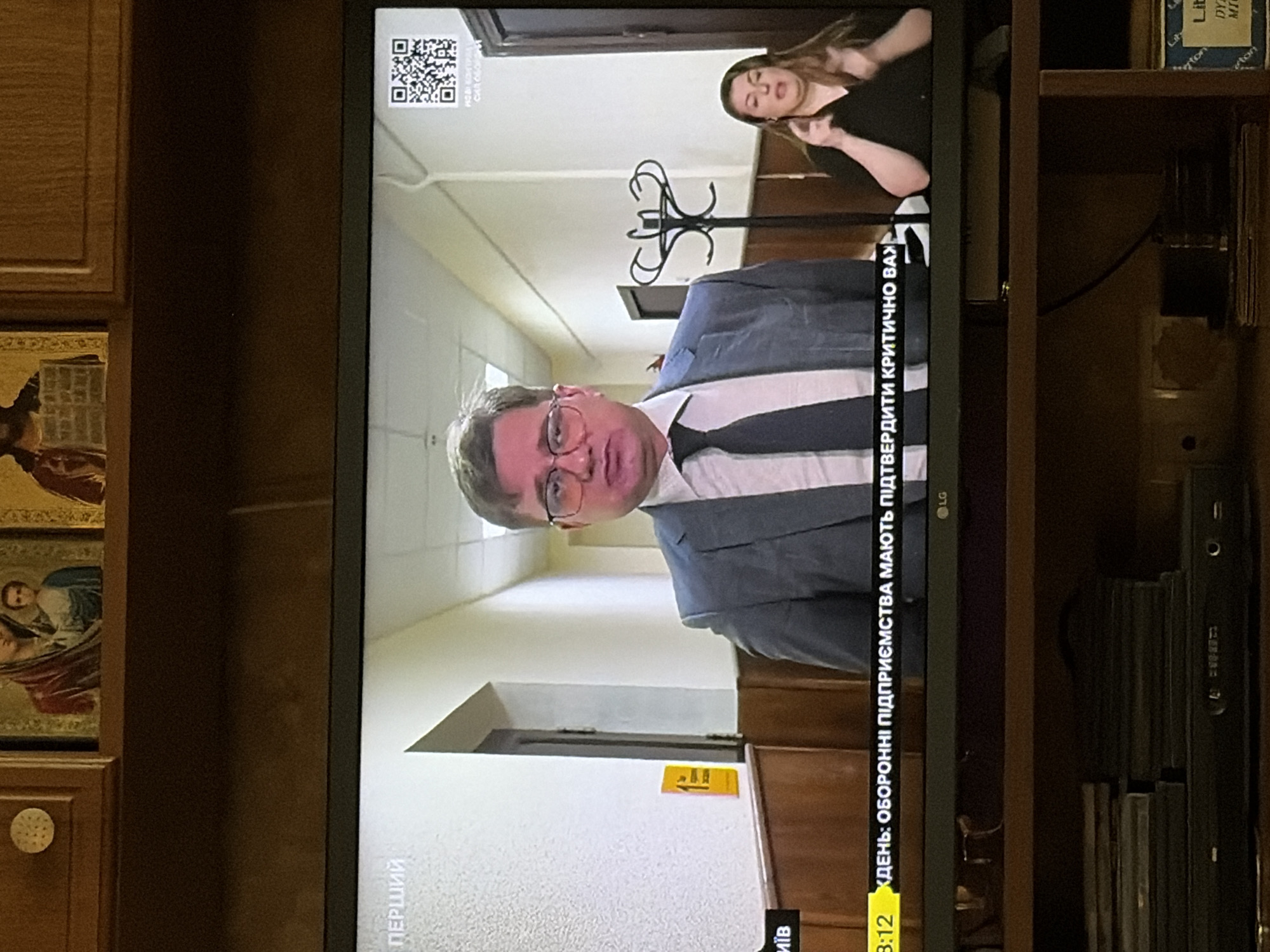 The image shows a television screen displaying a news broadcast. In the center of the screen is a man wearing glasses, a suit, and a tie, who appears to be a news anchor or correspondent. To his right, a woman is visible, likely a sign language interpreter. A news ticker is displayed at the bottom of the screen with text in Ukrainian. The background of the broadcast shows a hallway with wood paneling and a coat rack. The television is placed on a shelf in a room with religious icons visible on the wall above it.