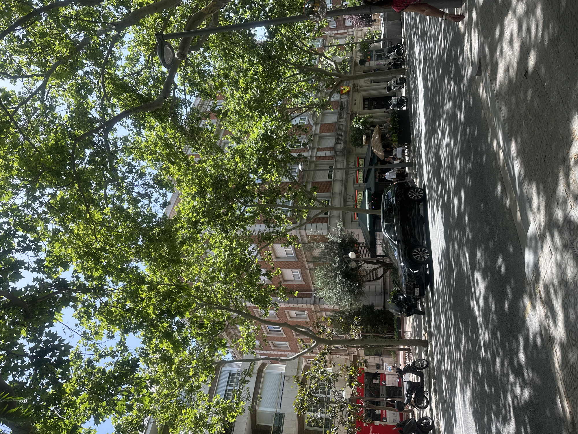 The image shows a sunny daytime scene on a city street, likely in Barcelona, Spain, with large trees casting dappled shadows across the pavement. A brick building with multiple stories and windows stands on the right side of the street, appearing to house businesses on the ground floor with awnings. In front of the building, there is a sidewalk cafe with tables and chairs. Several parked vehicles, including motorcycles and a black SUV, are visible on the street and sidewalk. A few people are present, some seated at the cafe and others walking. The overall atmosphere is that of a lively urban environment.
