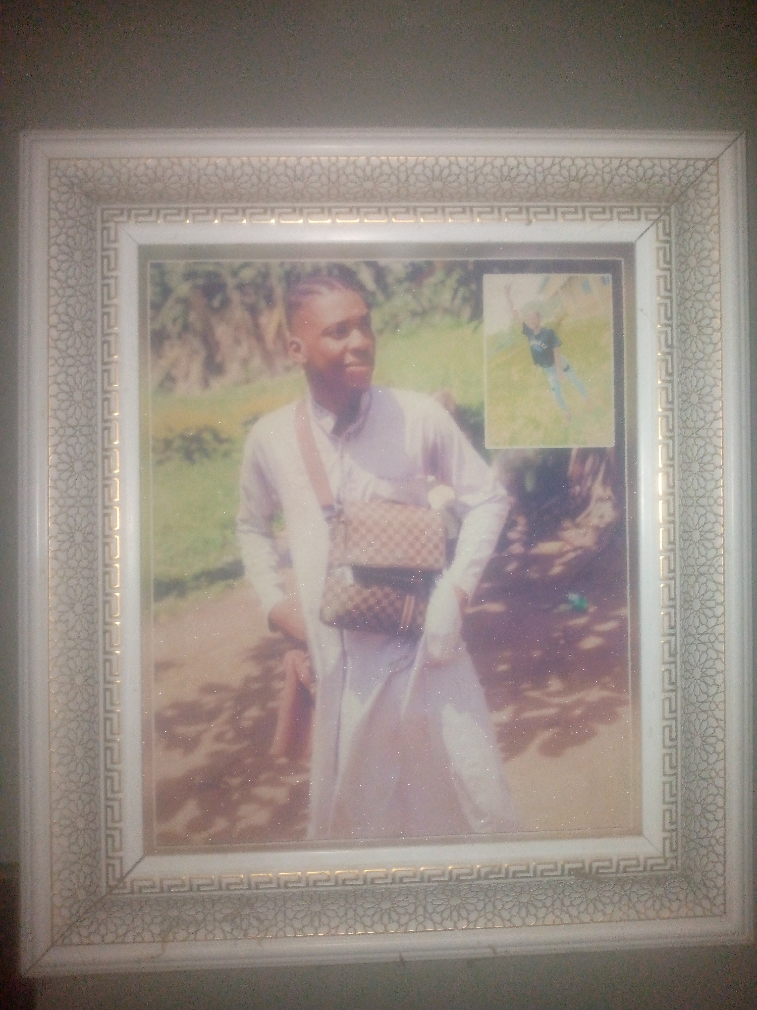 A framed photograph is centered in the image. The frame is white with a repeating gold Greek key pattern and a floral pattern around the edges. Inside the frame is a photo of a person with short braided hair wearing a long white shirt and a brown patterned cross-body bag. There is a smaller photo visible in the upper right corner of the larger photo, depicting a person in a dark shirt and blue jeans. The background of the main photo shows green foliage. The location of the city in the image is unknown.