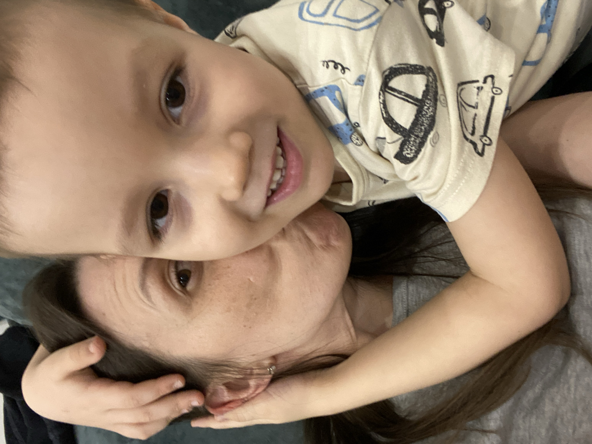 A young child lies on top of an adult. The child is wearing a light-colored shirt with vehicle designs. The child smiles with teeth showing. The adult has brown hair and light skin. The location is unknown and cannot be determined to be Chaikovsky, Russia.