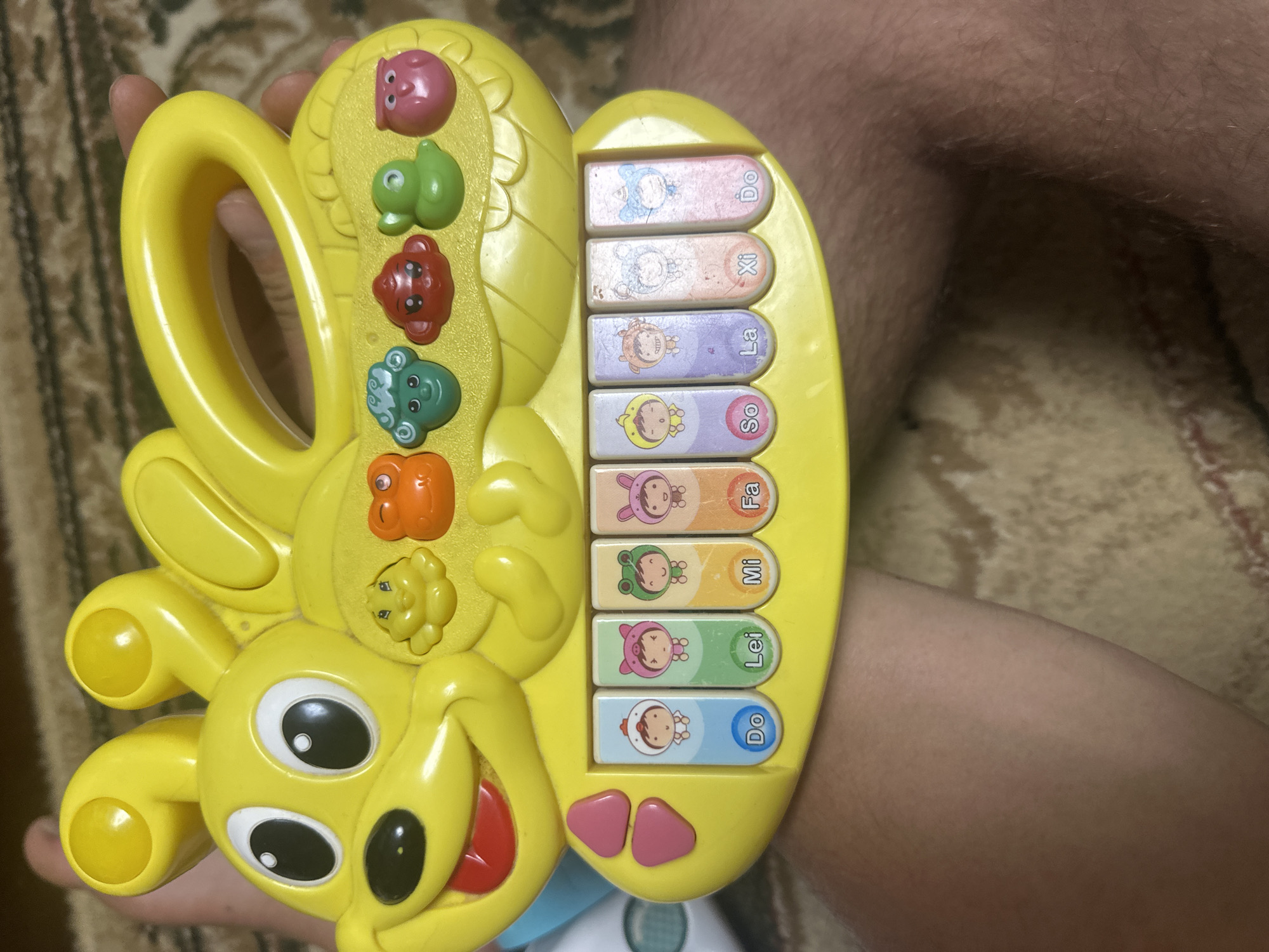 The image shows a close-up, eye-level view of a yellow, plastic toy keyboard in the shape of a cartoon caterpillar with rabbit ears. The toy is positioned on top of a person's lap, whose legs are visible at the bottom of the frame. The keyboard has several colorful buttons with images of cartoon characters, and musical notes labeled "Do," "Lei," "Mi," "Fa," "So," "La," "Xi," and "Do." Above the keyboard, there are several colorful buttons with animal faces. The background is a patterned rug, suggesting an indoor setting. The lighting is natural and appears to be from an overhead source. There is no visible indication of the city of Загородный, Russia, or any specific location within it; the image depicts a personal object and a person in an unidentifiable domestic environment.