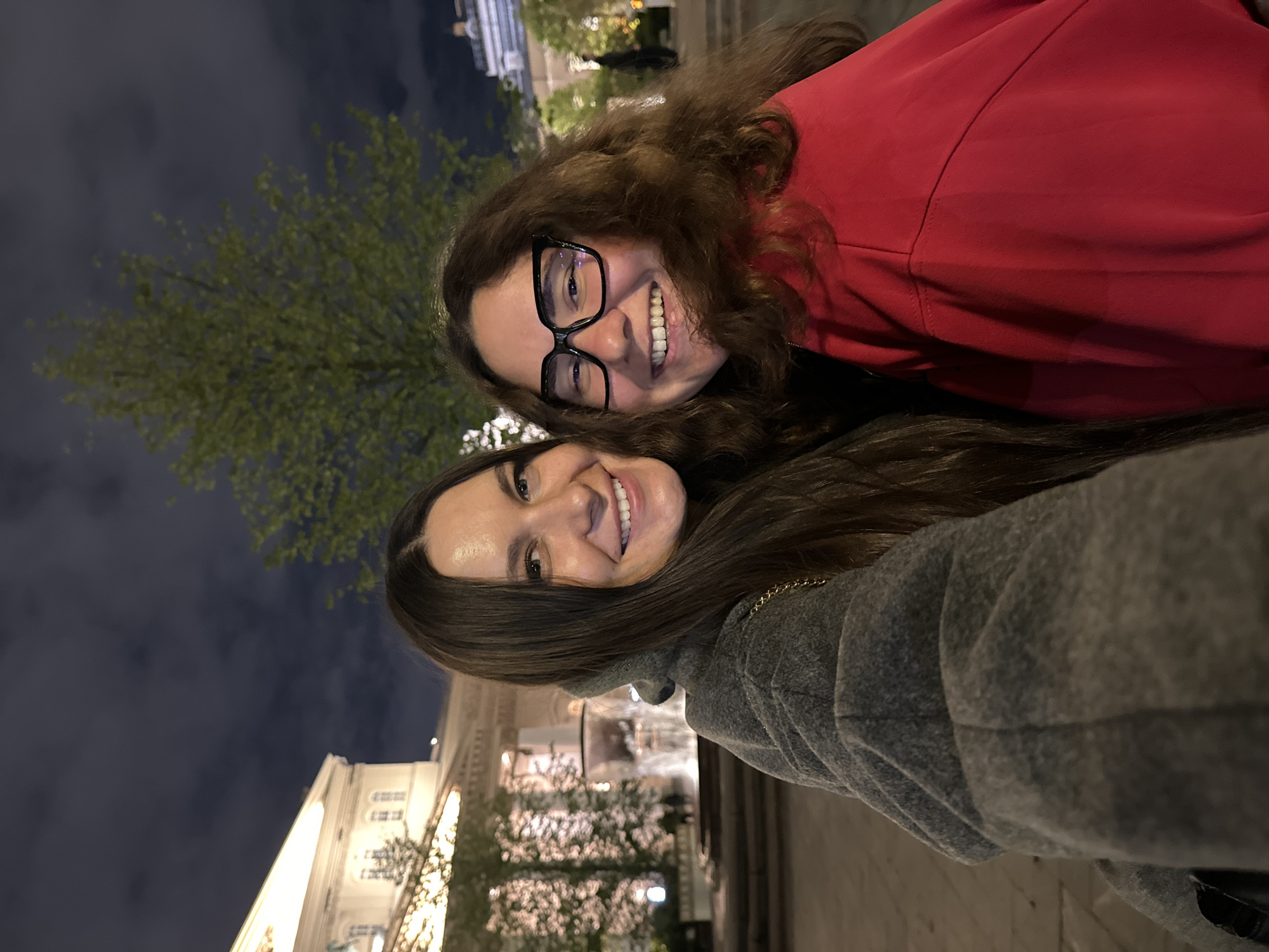 Two smiling women are posing for a selfie at night. The woman on the top wears glasses and a red jacket. The woman on the bottom wears a grey hooded sweater. Behind them are trees, the night sky, and a building illuminated with lights. Based on the architecture of the building, the location appears to be in Moscow, Russia, specifically near Red Square, where buildings with similar designs are found.