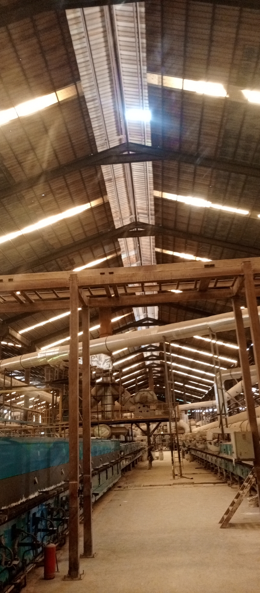 The image displays the interior of a large industrial facility or factory, likely located in Magboro, Nigeria. The perspective is looking down a wide central aisle, offering a view from the floor towards the high, vaulted roof.
The ceiling is constructed from corrugated metal sheeting, punctuated by numerous translucent skylights that allow abundant natural light to penetrate the space. These skylights create bright, almost glaring streaks of light across the roof structure, indicating it is daytime. Heavy metal beams and trusses support the roof.
Below the roof, an intricate network of industrial machinery and infrastructure is visible. Large, insulated pipes, possibly for conveying materials or air, run horizontally and vertically, supported by extensive metal frameworks and platforms. On the ground level, two long, parallel lines of industrial equipment define the central aisle. The machinery on the left consists of large, rectangular units primarily in blue-green, while those on the right are lighter in color. A red fire extinguisher is placed on the floor near the blue-green machinery on the left side. Various smaller pipes, electrical conduits, and support structures are integrated throughout the machinery. The floor is made of concrete and appears dusty.
No people are present in the image, and no specific activity or event is actively taking place, though the setup clearly indicates an operational industrial environment. There is no visible text discernible within the frame.