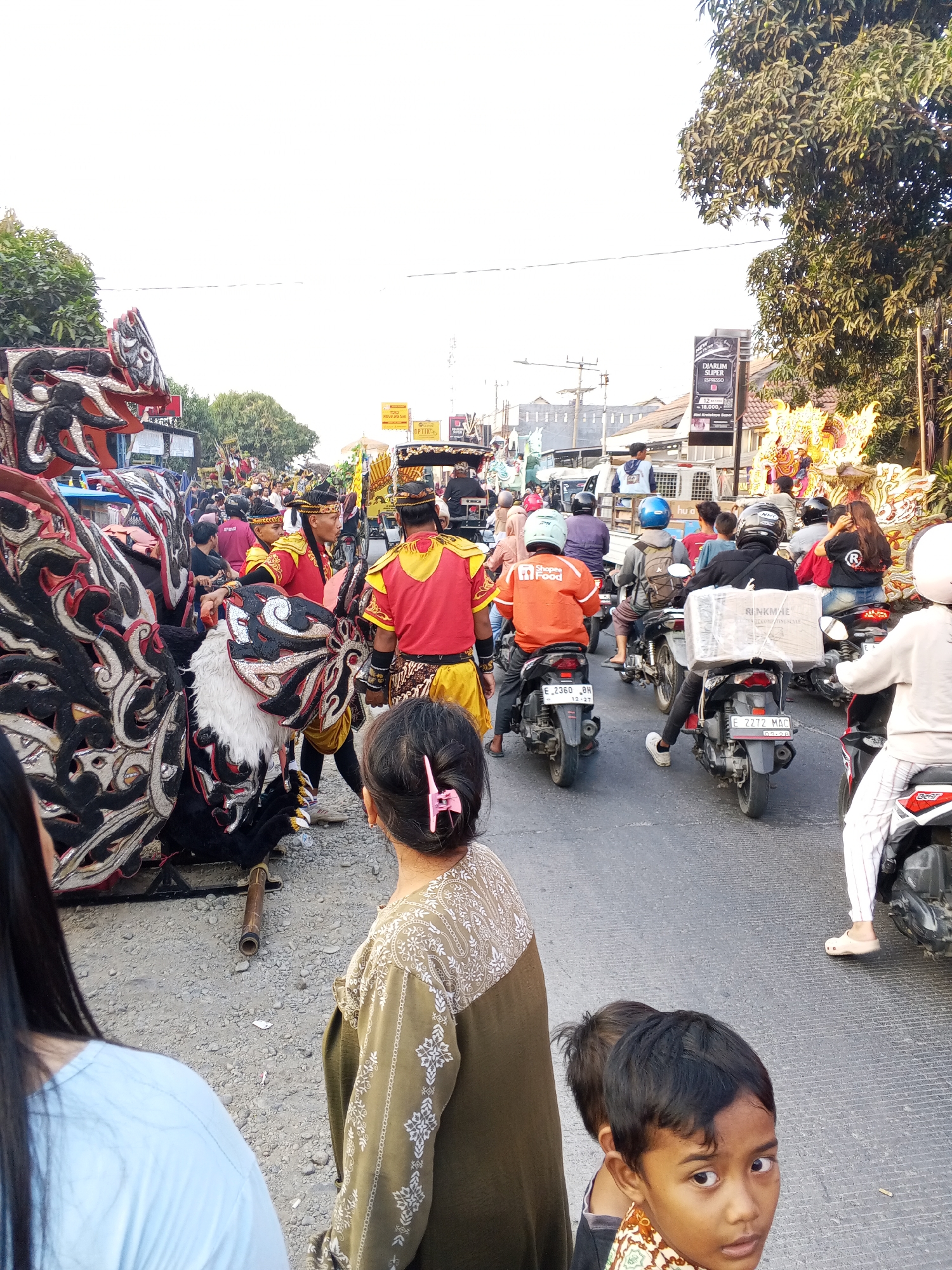 This image captures a street scene in Gebang, Indonesia, likely during a festival or parade. In the foreground, two children are visible, with one boy looking directly at the camera. Behind them, a woman in a patterned olive-green shirt stands, facing away. To the left, a large, elaborate puppet or costume, resembling a dragon or mythical creature, dominates the frame, adorned with black, white, and red patterns. Several people in traditional red and yellow costumes are near the puppet. The street is filled with motorcycles carrying riders and passengers, some of whom are wearing helmets. One motorcyclist wears an orange jacket with "Shopee Food" branding. In the background, more spectators and participants are present, along with buildings and trees lining the street. The atmosphere appears to be lively and crowded, suggesting a community event.
