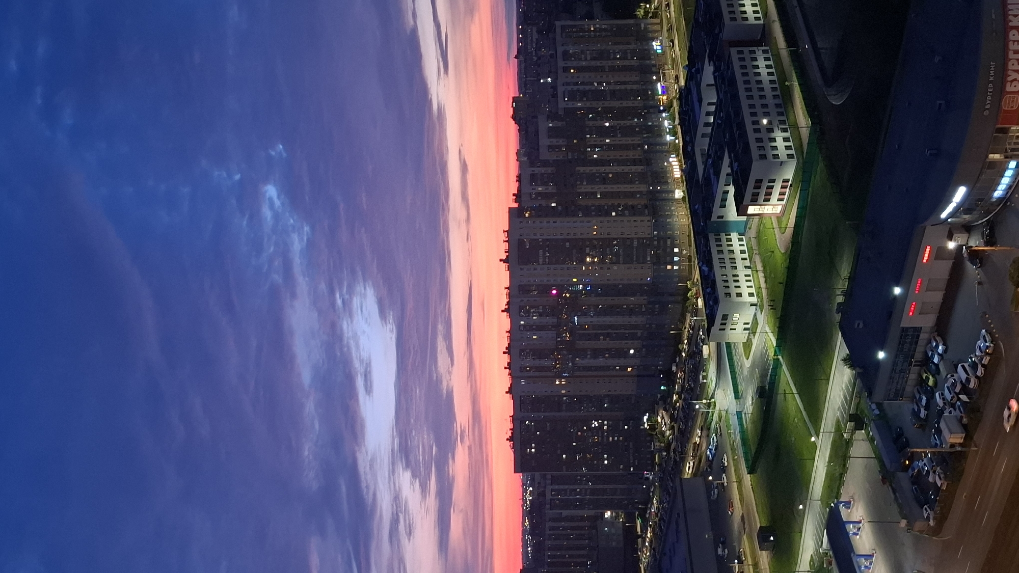 This is a vertical, high-angle shot of a modern cityscape at twilight, likely in an urban area of Russia, given the Cyrillic text visible on the buildings. The sky transitions from deep blue at the top to vibrant pink and orange hues near the horizon, indicating sunset or sunrise.
Dominating the mid-ground are several large, multi-story apartment buildings with many windows illuminated, suggesting occupancy. These buildings are tall and appear to be part of a recently developed residential complex. In front of these buildings, there are modern, low-rise structures with white facades and dark roofs, possibly educational institutions or administrative buildings. These are surrounded by green lawns and illuminated parking lots with numerous cars.
In the foreground, a dark, modern building with a curved facade features prominent, illuminated signage. The Cyrillic text on this sign includes "БУРГЕР КИНГ" (Burger King), identifying a fast-food restaurant. Adjacent to this building is a smaller, dark structure with what appears to be a gas station canopy. A road with visible car headlights and taillights runs along the bottom of the image. The overall impression is one of a well-lit, contemporary urban environment.