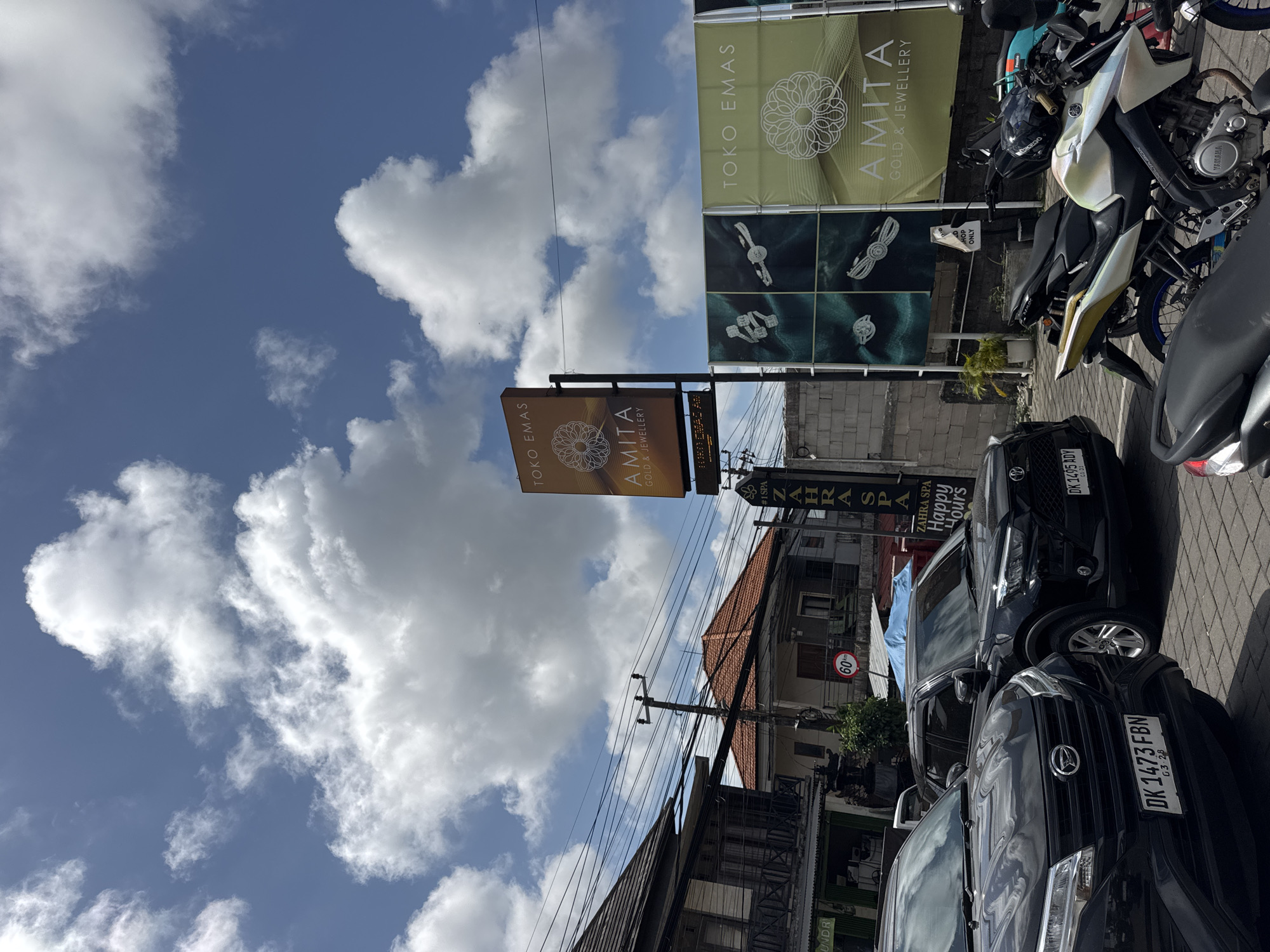 A street-level, wide-angle shot captures a sunny day in Kuta Selatan, Indonesia. The sky is a brilliant blue, dotted with fluffy white clouds. In the foreground, two black SUVs are parked on a cobblestone street. The one on the left has its front end visible, showing a license plate reading "DK 1473 FBN G3 28." The SUV on the right is also black, with a license plate that reads "DK 1495 ADY GI 21." The street is lined with buildings, including a two-story structure with a balcony and ornate railings on the left. A prominent orange sign with "TOKO EMAS AMITA GOLD & JEWELLERY" is visible, along with large green banners advertising jewellery. A vertical sign for "ZAHRA SPA Happy Hours" is also present. Wires are strung across the scene, supported by utility poles. In the background, more buildings with red-tiled roofs are visible. Several motorcycles are parked behind the SUVs. The overall impression is of a bustling commercial area on a clear day.