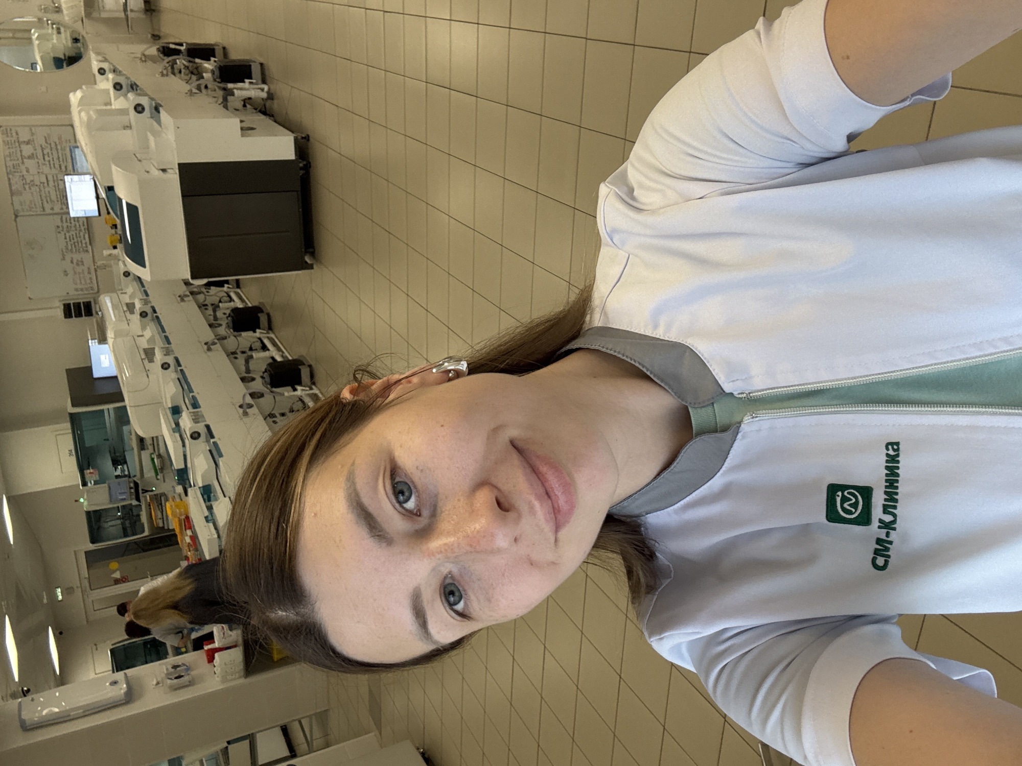 The image shows a young woman in a white lab coat with a green trim, standing in what appears to be a laboratory or medical facility. She is smiling and looking directly at the camera, with her hair down. She is wearing a light blue t-shirt underneath the lab coat. The lab coat has a logo and text on the left side of the chest, which reads "SM-Клиника" in green lettering, along with a green square logo containing a stylized "W" inside a circle.
In the background, there are rows of complex medical equipment, including what look like automated analyzers or laboratory instruments. The room has tiled walls and floors, typical of a sterile environment. The lighting suggests it is daytime indoors. The overall impression is of a professional setting, likely a hospital or a diagnostic laboratory in Moscow, Russia. The woman appears relaxed and friendly, possibly taking a selfie during a break or as part of an informal documentation.