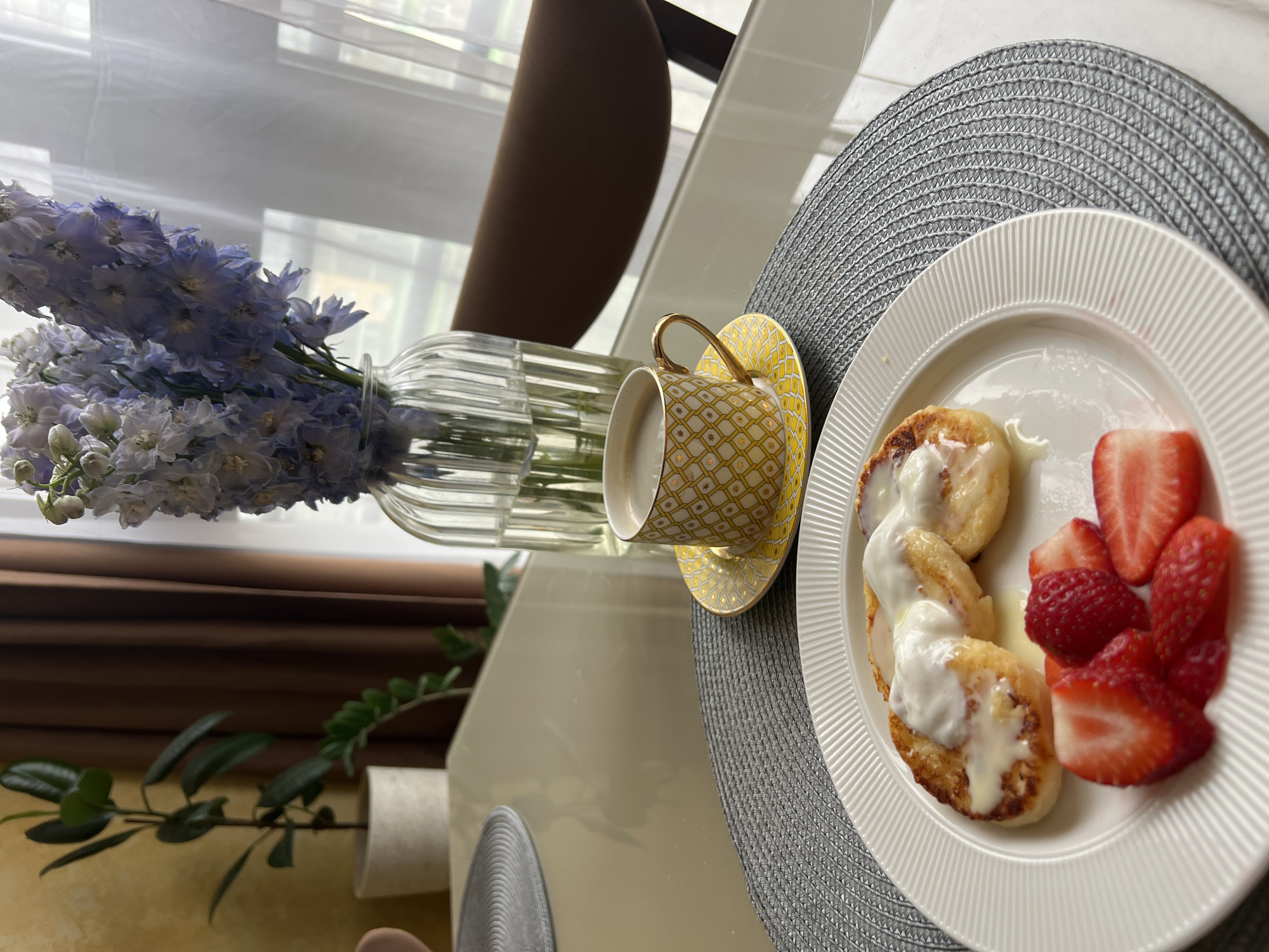 This image captures a bright and inviting meal setting in Khimki, Russia, likely during morning or early afternoon due to the soft, natural lighting. The scene focuses on a table laden with breakfast or brunch items.
In the foreground, a white, subtly ridged plate holds three golden-brown, pan-fried cheese pancakes (syrniki) generously drizzled with a creamy white sauce, possibly sour cream or condensed milk. Alongside them, a vibrant cluster of fresh red fruit includes both whole raspberries and sliced strawberries.
To the left of the plate, a small, elegant teacup with a gold handle and a delicate yellow and gold geometric pattern sits on a matching saucer, filled with a light-colored beverage, possibly coffee with milk or a latte. Both the cup and plate rest on a round, textured dark grey placemat.
A decorative element, a clear, ribbed glass vase, holds a beautiful bouquet of light blue to lavender flowers, possibly delphiniums, adding a touch of natural beauty to the arrangement. The table itself is a shiny, light-colored surface that reflects parts of the surrounding objects.
The background is softly blurred but hints at an indoor environment with white blinds or curtains, a dark brown vertical element (perhaps furniture or a window frame), and some green foliage from an indoor plant. No people are visible, and there is no discernible text on any of the items. The overall impression is one of a calm and pleasant meal.