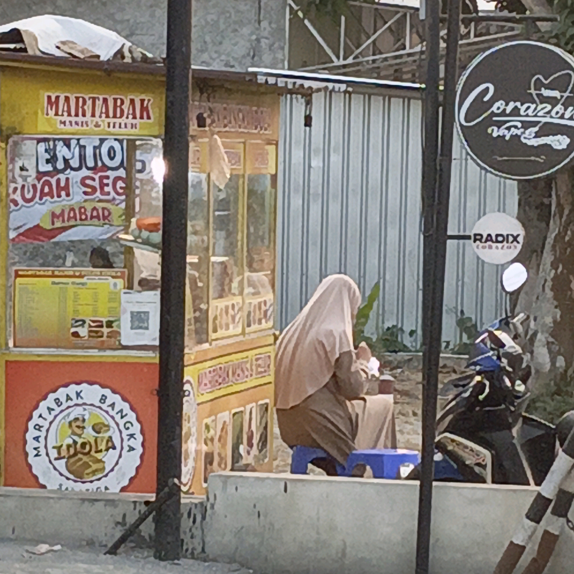 This is a medium shot, taken at an eye-level angle, of a street food stall in Gedangan, Indonesia. The stall is yellow and has red and white signage. One sign reads "MARTABAK MANIS & TELUR". Another sign reads "ENTONG RUAH SEGER MABAR". To the left of the stall, a circular logo displays a chef and the text "MARTABAK BANGKA IDOLA".
A person wearing a light brown hijab and a brown garment is seated on a small blue stool in front of the stall. They are facing away from the camera and appear to be preparing food.
To the right of the stall, a motorcycle is parked. A circular sign with white lettering on a dark background reads "Corazon Vape". Another circular sign below it reads "RADIX TOBACCO".
The environment appears to be an outdoor setting, possibly a street or marketplace. The background includes a corrugated metal wall and some trees. The lighting suggests it is daytime, with some shadows present. A concrete barrier runs along the bottom of the frame, with some litter on it.