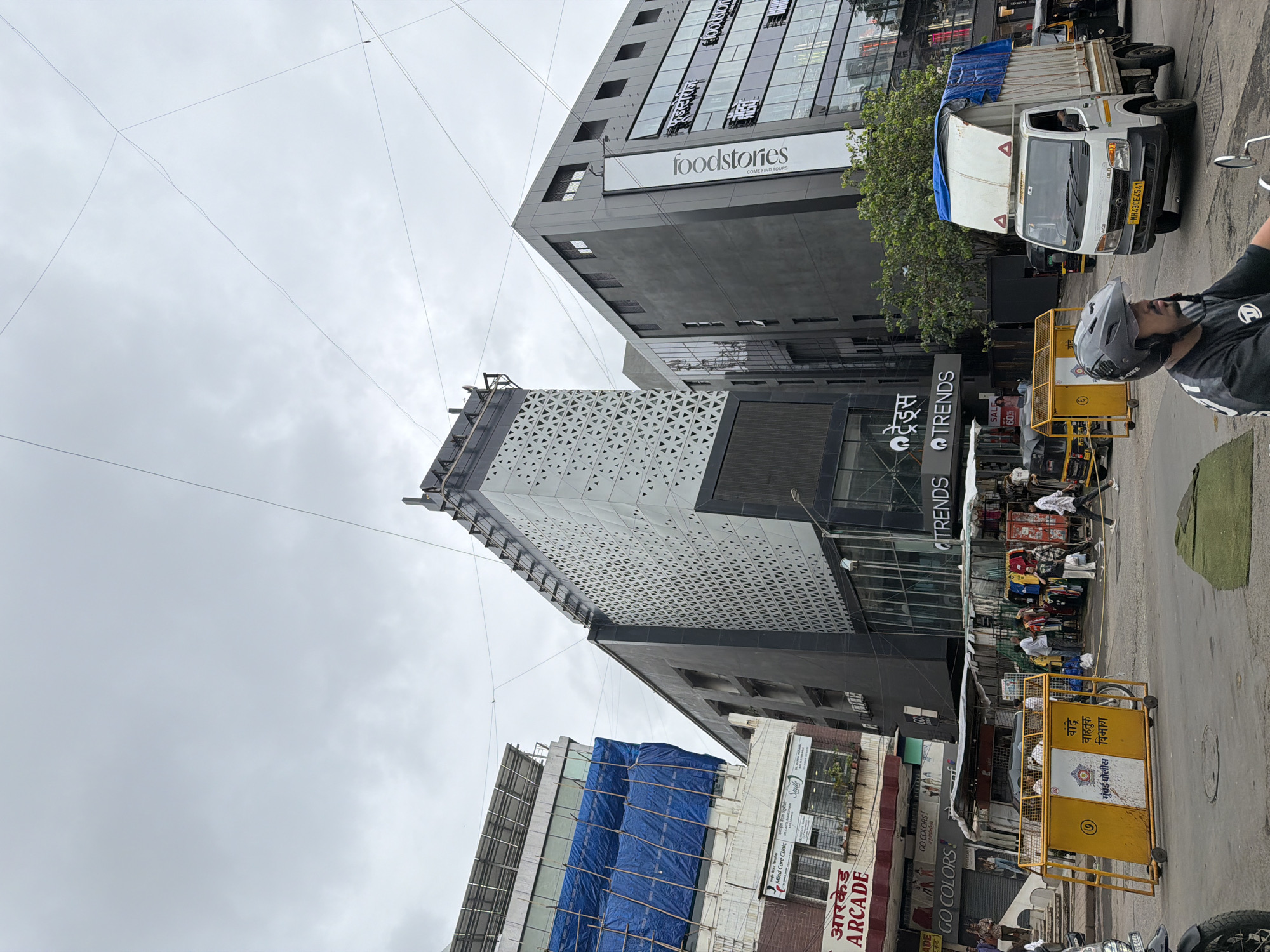 The image depicts a street scene in Mumbai, India, on an overcast day. In the foreground, a man wearing a black helmet is riding a motorcycle. To his left, a yellow police barricade with Marathi text and a Mumbai police emblem is visible. Further down the street, various shops are open, with awnings and displays of clothing. A white truck covered with a blue tarp is parked on the right side of the road, alongside an auto-rickshaw. In the background, several buildings are visible, including a modern building with a white patterned facade and a darker building with the "foodstories" logo. Signs for "TRENDS," "ARÇADE," and "GO COLORS!" are also present. The sky is cloudy, and power lines crisscross the upper portion of the image.