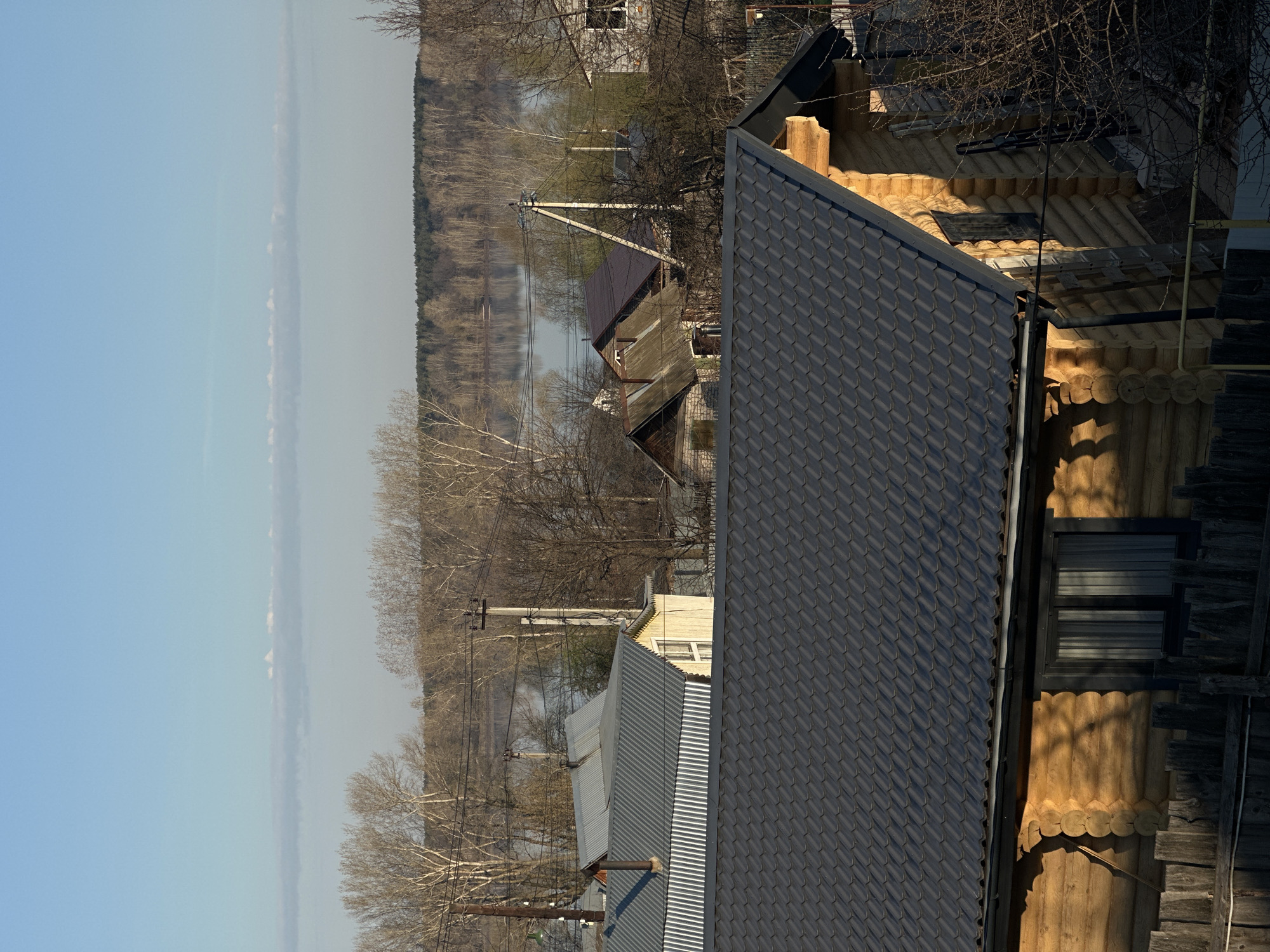 The image shows an aerial view of a residential area in Chaikovsky, Russia. The scene features a mix of buildings, trees, and a body of water.
Dominating the foreground is a house with a dark gray shingled roof. Adjacent to it is a log cabin-style building with exposed round logs and a window. Behind these structures, there's another house with a gray corrugated metal roof and a yellow facade.
In the background, a body of water is visible, possibly a river or lake, with a forest lining the opposite bank. Bare trees and utility poles dot the landscape in the middle ground, adding to the urban feel. The sky is light blue.