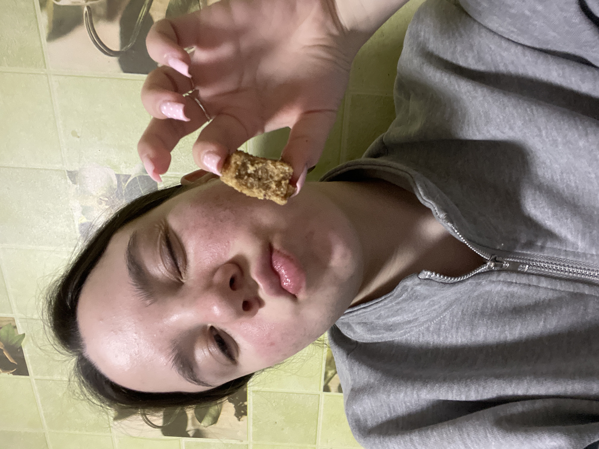 A young person with light skin, closed eyes, and dark hair is holding a small, roughly rectangular piece of food between their thumb and forefinger. The person is wearing a gray zippered sweatshirt. The background appears to be a kitchen with tiled walls. No information in the image suggests this is in Ольховка, Russia.