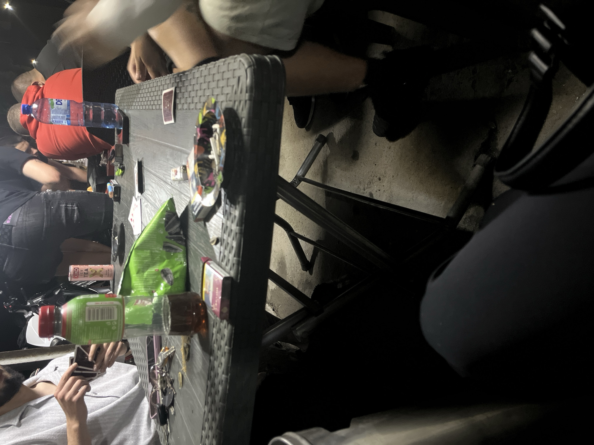 The image depicts a group of individuals gathered around a table outdoors, likely in the city of Plovdiv, Bulgaria. The setting appears casual, possibly a backyard or a public seating area. On the table, various items are visible: a bottle of water, a green bottle of what appears to be tea, a packet of chips, a pack of cigarettes, an ashtray with cigarette butts, playing cards, and a mobile phone.
The individuals are partially visible, with their focus seemingly on the table and activities at hand. One person on the left is holding playing cards, suggesting a game is in progress. Another person in the center is wearing a red hoodie, and another on the right is wearing dark clothing. The overall lighting suggests it is either evening or night. The ground beneath the table is concrete.