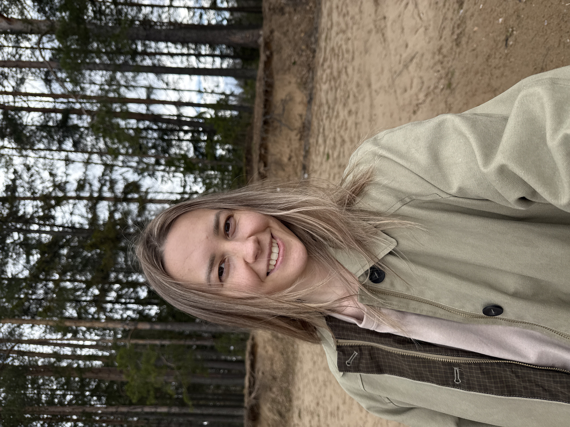This is a bright outdoor portrait of a smiling woman in a forest setting, located in Chaikovsky, Russia. The woman, with light brown to blonde hair, is positioned in the foreground, facing the camera with a wide, joyful smile that shows her teeth. She wears a light olive-green casual jacket with dark buttons and a checkered brown interior lining, over a pale pink or beige top. Behind her, a dense forest of tall, slender coniferous trees, likely pines or spruces, stretches upwards, with patches of bright sky visible through their branches. The ground appears to be a sandy or light brown dirt embankment, visible to her side and behind. The scene is illuminated by natural daylight, suggesting a daytime photograph taken in possibly cool or mild weather. No specific activity beyond posing for the photograph is evident, and there is no visible text.