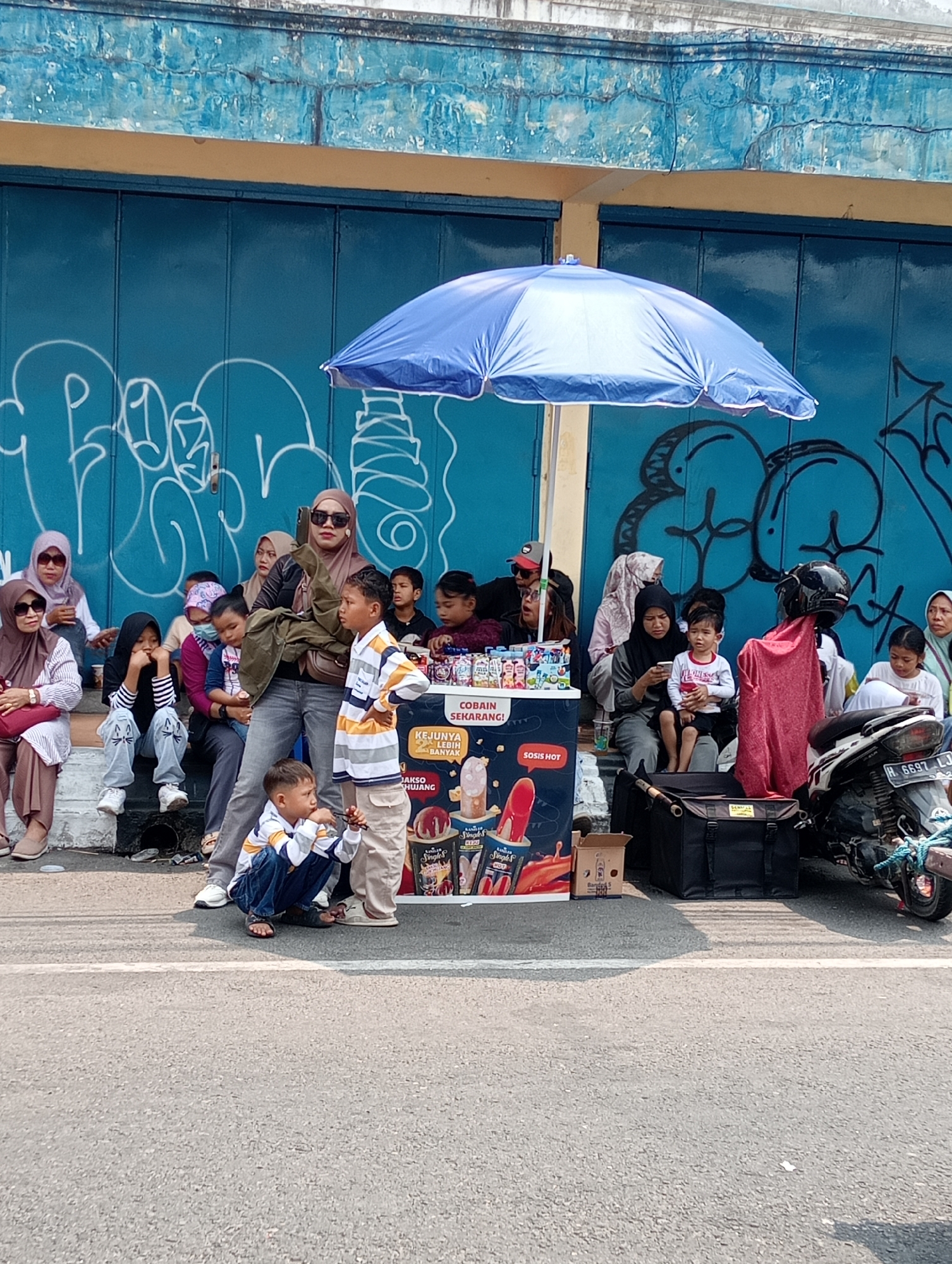 The image depicts a street scene in Demak, Indonesia, likely near a marketplace or a busy thoroughfare, given the presence of a food cart and various people. A blue awning shades a cart that appears to be selling ice cream and hot dogs, with colorful packaging visible. A group of people, including adults and children, are gathered around the cart and sitting on a low wall or steps behind it. The background features a blue-painted wall with graffiti. A motorcycle is parked to the right of the frame. The overall impression is one of a casual, everyday gathering in a public space.