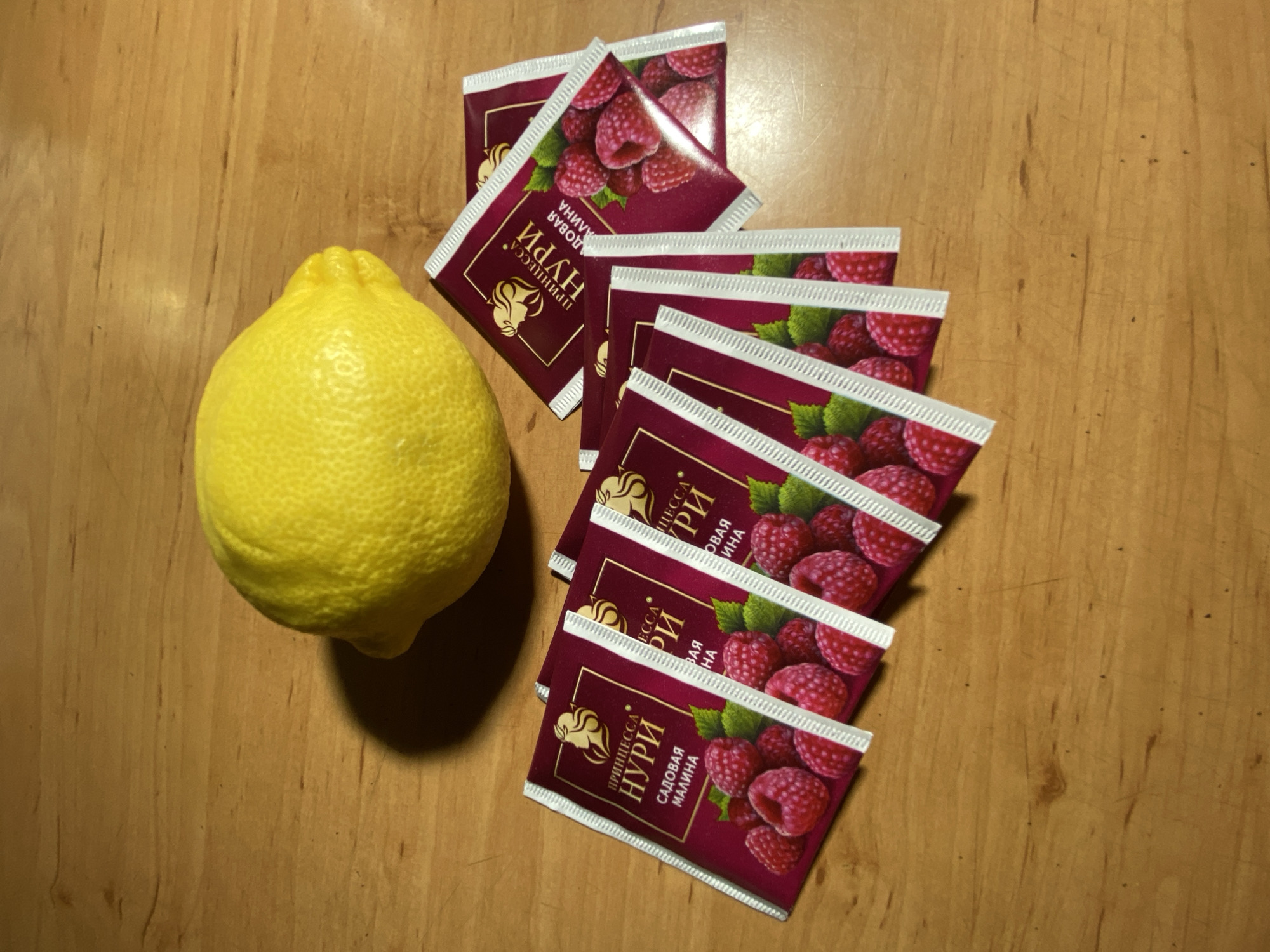A single bright yellow lemon rests on a wooden surface next to a fan of several small, individually wrapped packets of raspberry-flavored tea. The packets are a deep burgundy color with a gold logo and an image of raspberries and green leaves. The tea packets are arranged in a fanned-out pattern, overlapping each other. The wooden surface appears to be a table with visible grain and some minor imperfections. The lighting suggests an indoor setting, with a shadow cast by the lemon. There is no indication of the city of Chișinău, Moldova, or any people present in the image.