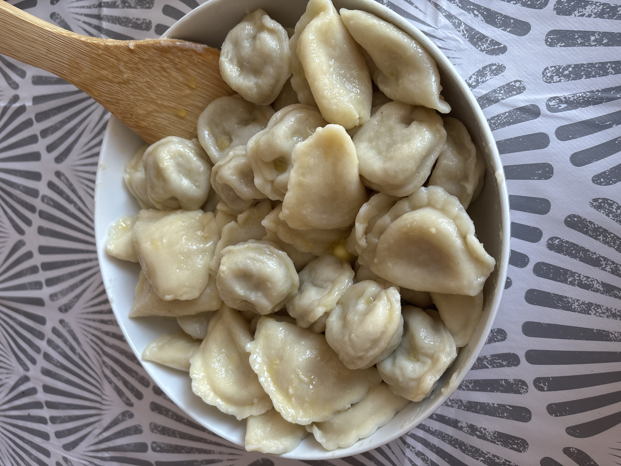 A white bowl filled with dumplings is placed on a table with a grey and white patterned tablecloth. A wooden spatula rests on the right side of the bowl, partially submerged in the dumplings. The dumplings appear to be cooked and glistening, suggesting they are either buttered or in a sauce. The lighting is bright, and the image is a close-up shot, focusing entirely on the food and the bowl. The scene is indoors. There is no information or visual evidence to determine the location in the city of Cork, Ireland.