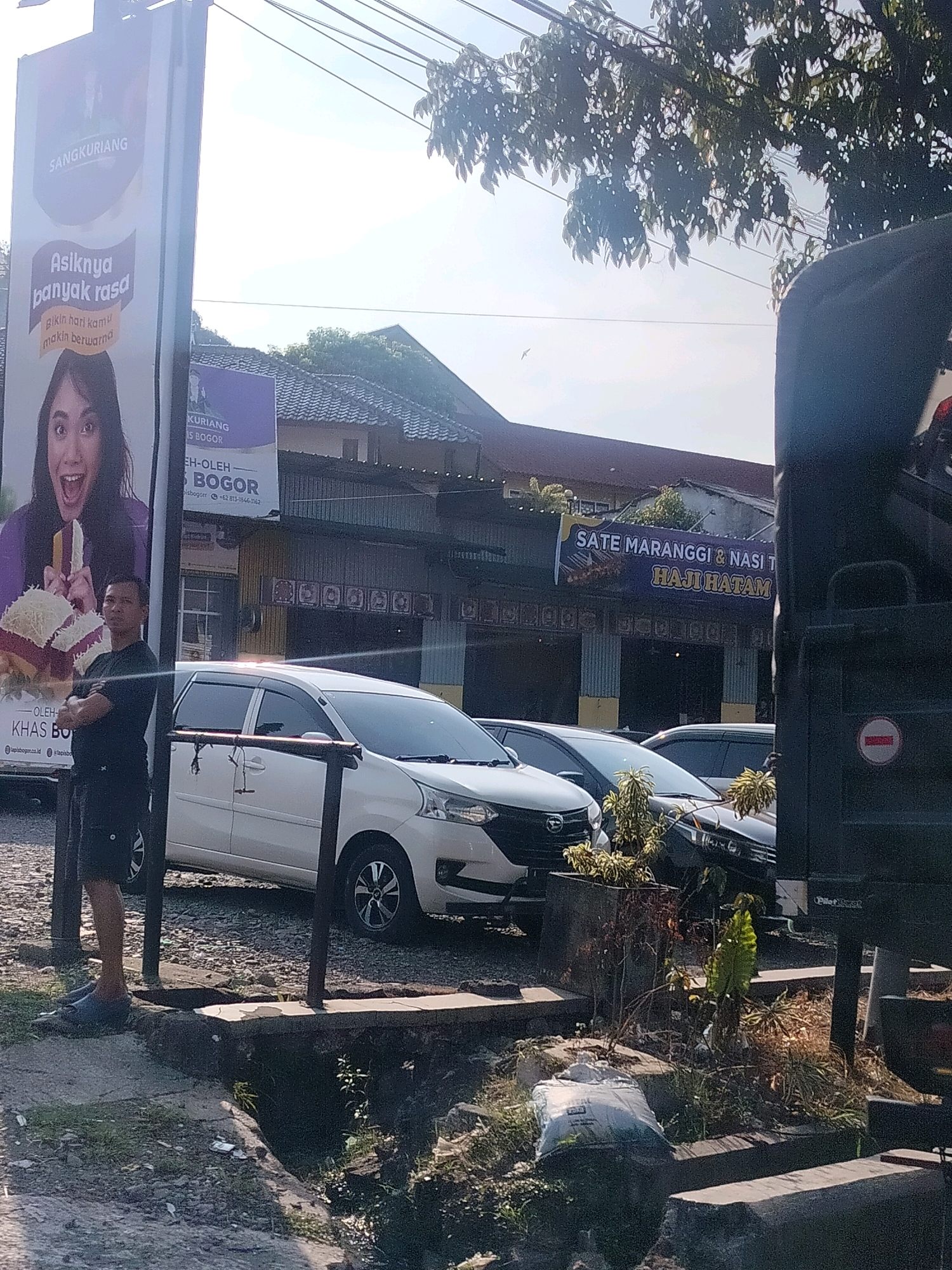 A man stands with his arms crossed in front of a large billboard advertising "SANGKURIANG Asiknya banyak rasa". Behind him, a white van and several other cars are parked in front of a row of buildings. One of the buildings has a banner that reads "SATE MARANGGI & NASI T HAJI HATAM". The scene appears to be in a street or parking area, possibly in Cisarua, Indonesia, given the context provided. The lighting suggests it is daytime.