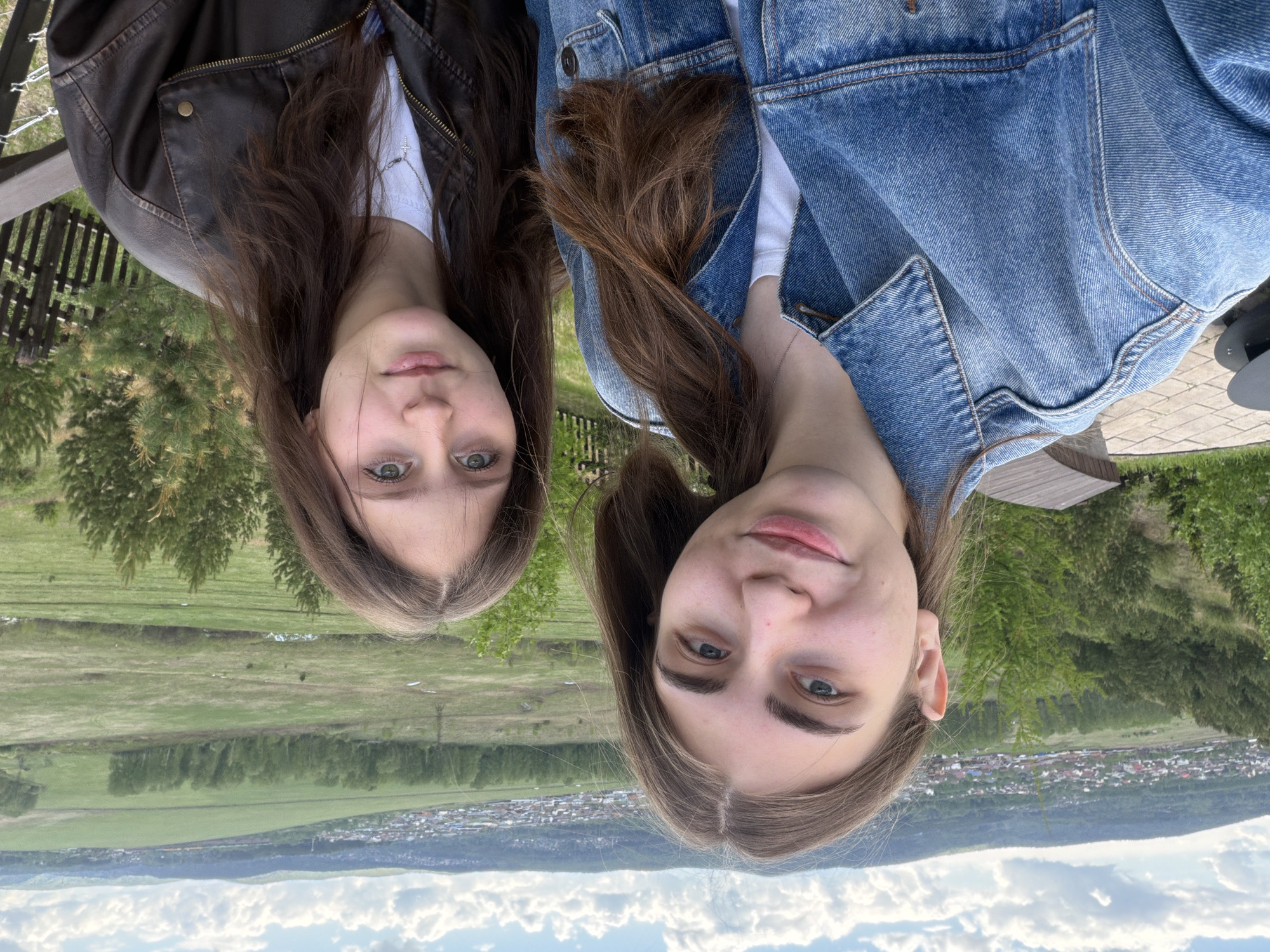 Two young women are seen from a high vantage point, appearing to be on a hillside overlooking a town and landscape. One woman is wearing a brown leather jacket over a white top and a necklace. The other is wearing a blue denim jacket over a white top with a necklace. The background consists of a green landscape with forests and a town nestled in a valley, with the sky visible in the distance. The city of Shamoni, Russia is not discernible from the image.
