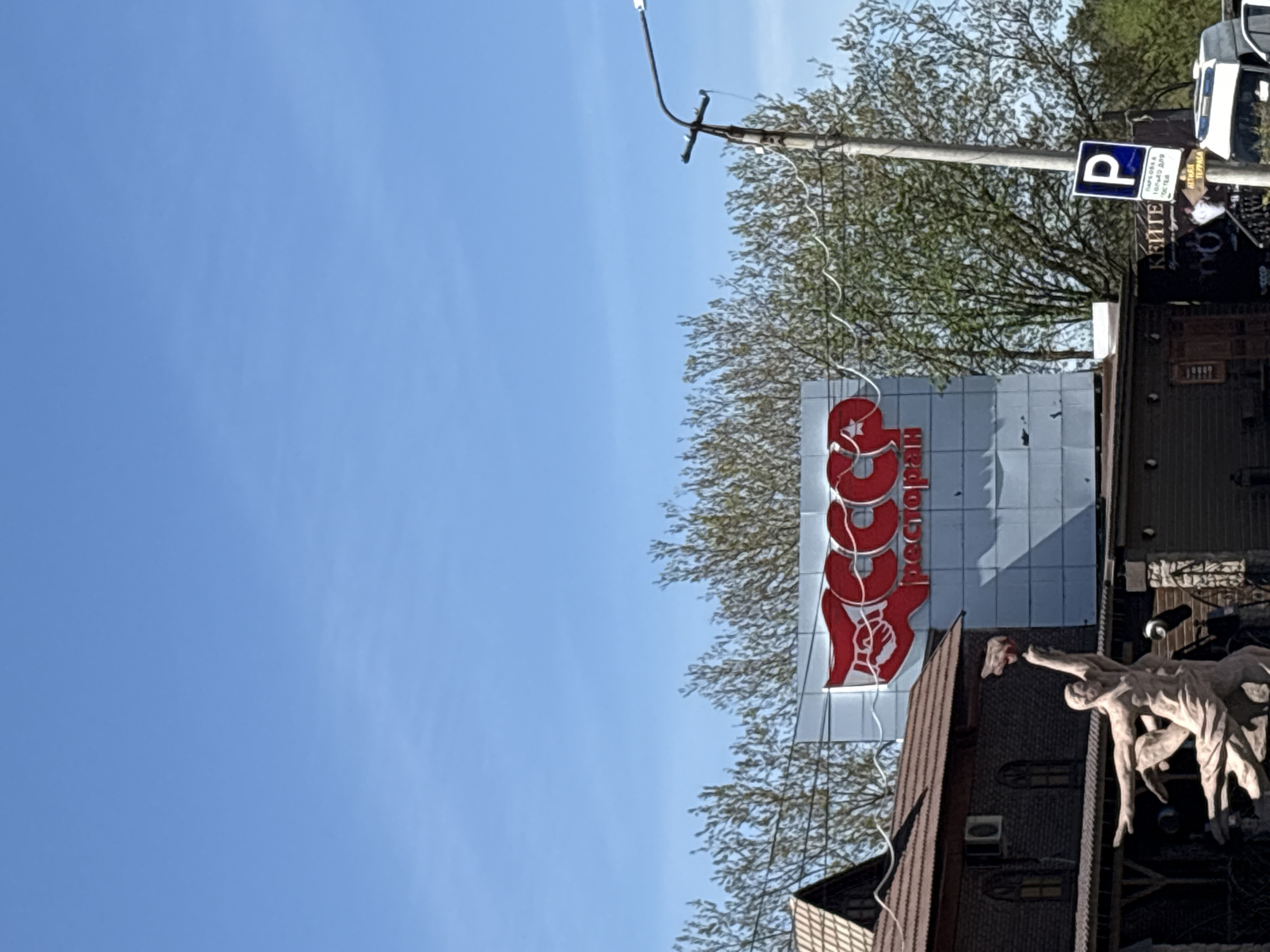 A vertical, eye-level shot depicts a clear blue sky occupying the left half of the image. The right half reveals a multi-story building adorned with a large sign featuring the word "СССР" (USSR) in red Cyrillic letters, indicating a restaurant. Below the sign, a sculpture of figures clings to the building’s facade. A power pole with wires runs above the building. Also visible is a parking sign affixed to the pole. This scene is located in Perm, Russia.