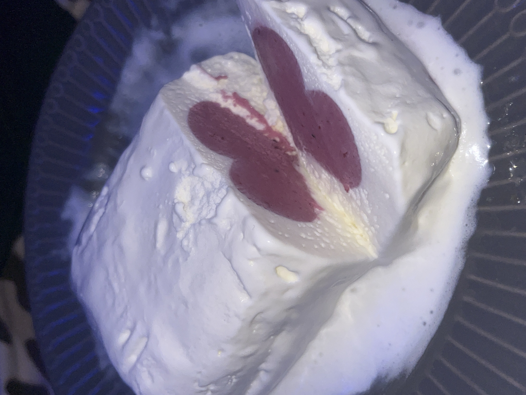 A close-up, high-angle shot captures a dessert, likely ice cream or a similar frozen treat, presented in a dark-colored bowl with visible ridged patterns. The dessert is predominantly white and appears to be melting slightly, creating a foamy, liquid substance around its base and edges.
The central subject is a portion of the dessert that has been cut, revealing its internal structure. It consists of a thick, creamy white outer layer, which seems soft and somewhat aerated. Within this white layer, there is a striking red or deep pink, fruit-flavored core or filling. This inner layer is distinctively shaped, appearing in two main sections that resemble elongated, organic blobs or perhaps abstract butterfly wings, separated by a thin strip of the white or a light yellow layer. The texture of the red/pink part seems smooth.
The melting process is evident from the liquid accumulation in the bowl and the slightly glossy appearance of the dessert's surface. The lighting is somewhat dim, casting shadows and highlighting the texture of the white dessert and the ridges of the bowl. No people are visible, nor is any discernible text. The setting appears to be indoors, focusing entirely on the dessert presentation.