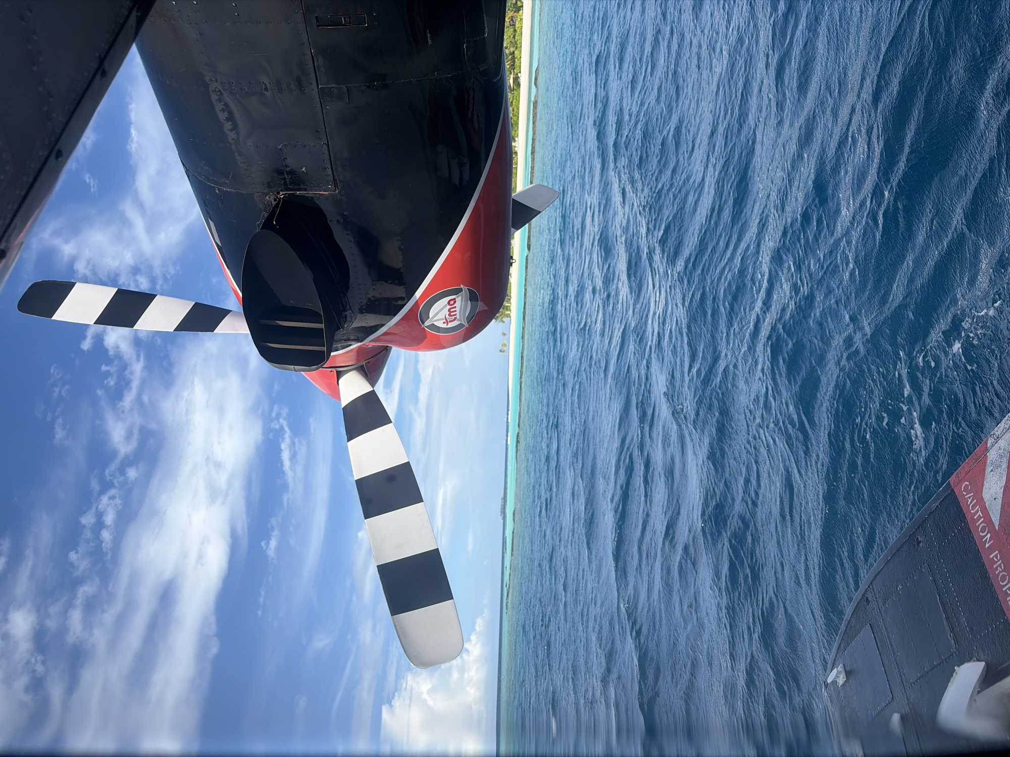 An overhead view of a Twin Otter seaplane's engine nacelle, propeller, and a section of its fuselage as it flies over the ocean. The propeller is black and white striped, and the nacelle is black with red accents and a logo that reads "tma". The ocean is a deep blue with small waves. In the background, a white sandbar with palm trees and a few small boats is visible. The sky is blue with scattered white clouds. The image was taken from inside the aircraft looking out. The location is over the water near Halaveli, Maldives.