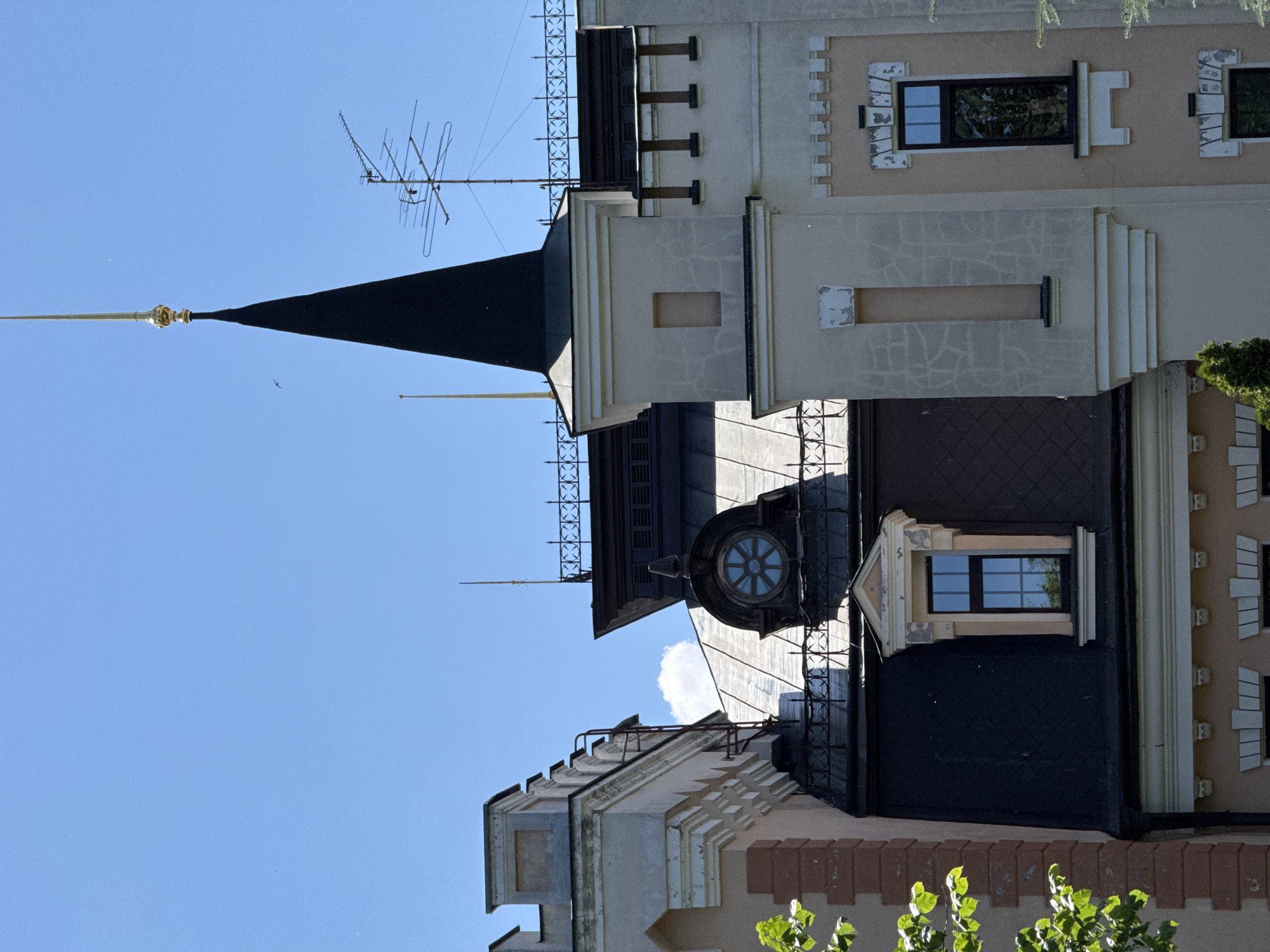This is a low-angle, full shot of the roofline of a building in Kyiv, Ukraine, on a clear day. The building features a prominent black, conical spire topped with a gold finial. To the right of the spire, an old-fashioned television antenna is visible. The building's facade is light beige, with decorative elements and a dark-tiled mansard roof. A small circular window with a distinctive blue, star-like pattern is set into the mansard roof. On the left side of the image, the corner of another building section is visible, with lighter colored stone and reddish-brown brickwork. Green foliage from a tree is present in the lower left corner, and a few small white clouds are in the bright blue sky.