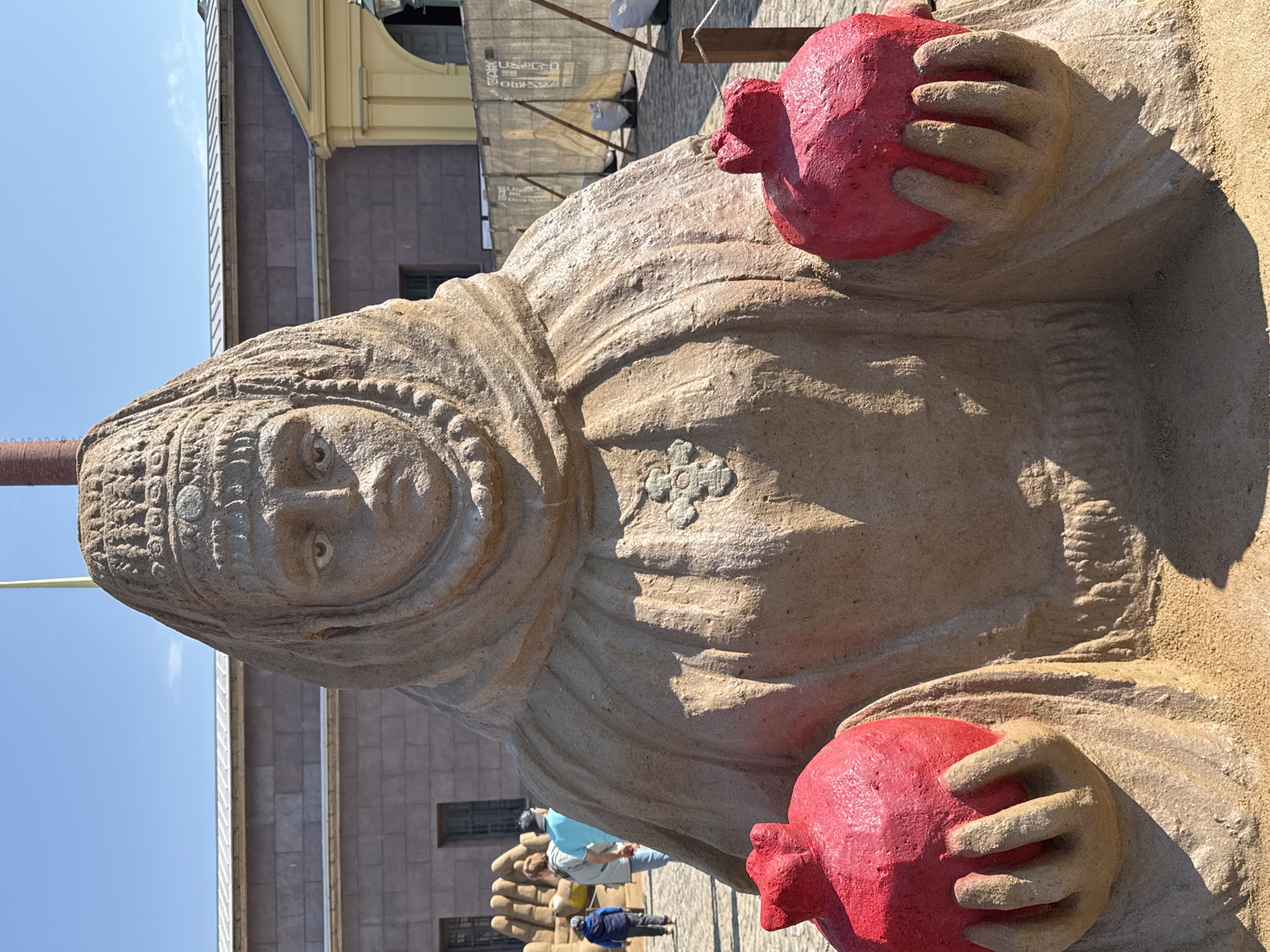 The image depicts a close-up, medium shot of a sandstone statue of a woman holding a red pomegranate in each hand. The woman is wearing a head covering with decorative edging and a simple gown with a cross-shaped embellishment on the chest area. Her facial expression is neutral, and the details of the statue show signs of weathering. The background includes a section of a building with visible windows, an area with scaffolding, and a blue sky visible in the upper left corner. Other statues are visible in the background and there are some people in the background as well. The location is the Peter and Paul Fortress, Saint Petersburg, Russia.