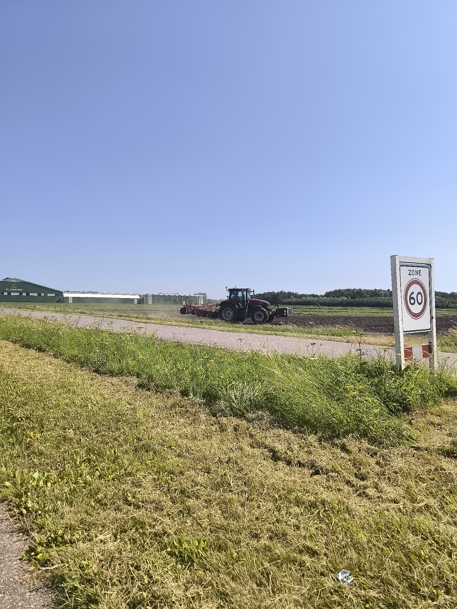 In Andijk, Netherlands, on a clear day, a red tractor is plowing a field near a farm. A speed limit sign indicating "ZONE 60" is visible in the foreground, suggesting a controlled speed area. The environment includes a paved road, a grassy verge, and agricultural buildings in the background. The sky is clear and blue.