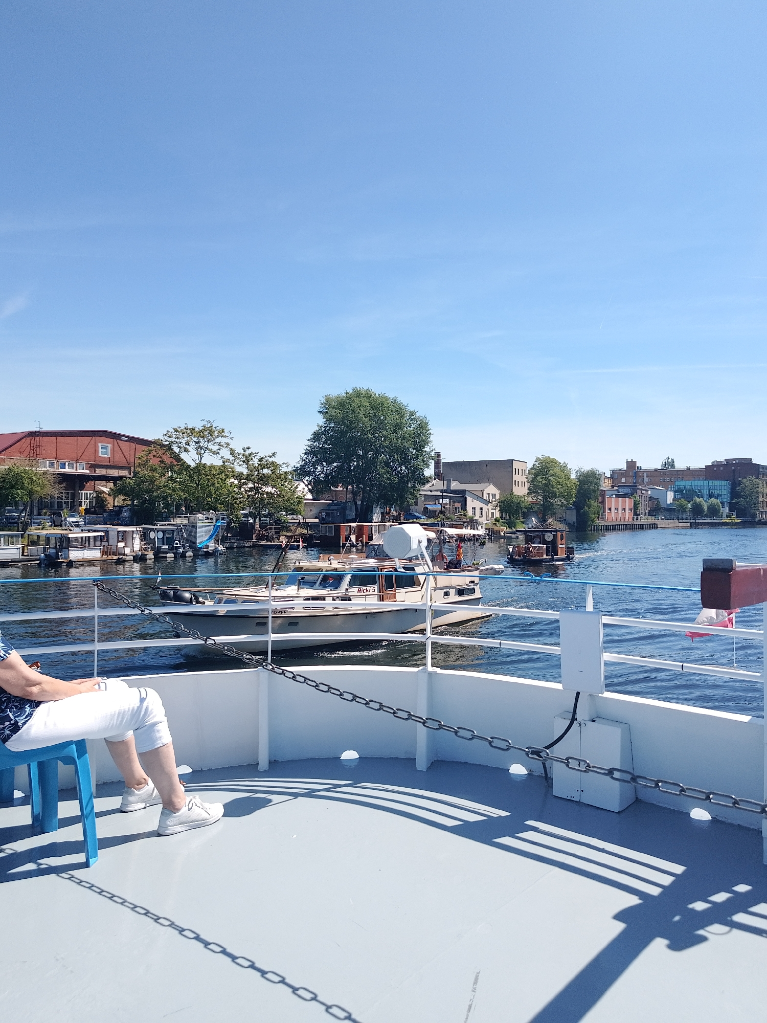 A medium-angle shot reveals a serene scene on a boat deck in an urban setting. A person is seated on a blue plastic chair to the left, wearing a blue patterned top and white pants with white sneakers. The deck's light gray surface is adorned with the shadows of the boat's railing. A white boat with the name "Ricki 5" is seen on the water. The cityscape in the background includes a variety of buildings, including red-brick structures, light-colored buildings, and a few modern glass-fronted buildings. The water surface reflects the blue sky. The location is in the city of Unknown, Unknown.