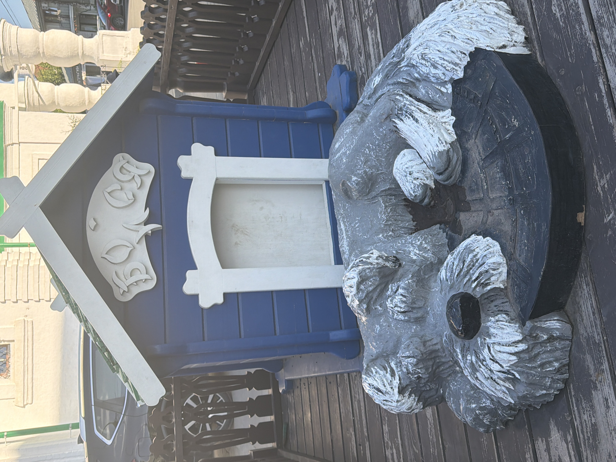 A weathered, grey, sculptural representation of a dog is positioned in front of a small, blue, wooden doghouse with white trim. The dog sculpture is curled up, appearing to sleep, with its head resting on a black, circular base. Its fur is textured and painted with white highlights. The doghouse has a decorative white carving on its gable depicting a stylized animal or face. The scene is set on a wooden deck, with a dark brown wooden railing to the right of the doghouse. In the background, a dark-colored car is partially visible behind the doghouse. Further in the background, architectural elements suggest a building with columns and windows, and other buildings, indicative of an urban or town setting. The lighting suggests it is daytime, with sunlight casting shadows. This appears to be a decorative element at a public location in Vologda, Russia, possibly a park or an establishment featuring this unique sculpture.