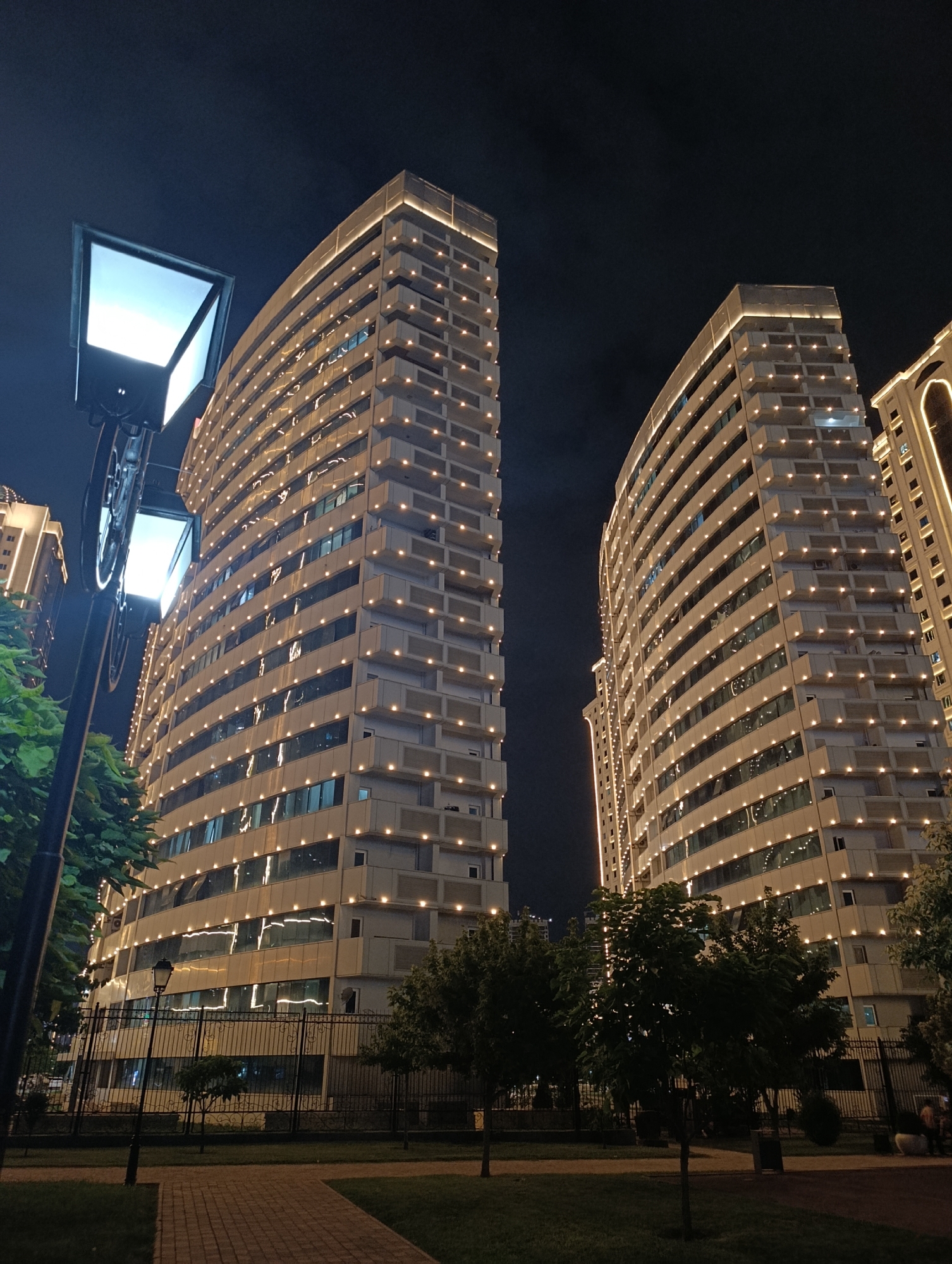 The image depicts a night view of modern apartment buildings in Grozny, Russia, specifically the Grozny City complex. Two tall, illuminated buildings dominate the scene, their facades covered in small, warm-toned lights that trace the architectural lines. The building on the left is curved and appears to have balconies or recessed windows. The building on the right is straighter and also features extensive lighting.
In the foreground, a street lamp with a rectangular illuminated panel stands to the left. The lamp is on a pole that also supports what appears to be a decorative metal wheel. Green trees and foliage are visible in front of the buildings and along a dark metal fence that encloses the area. A brick pathway leads through a grassy area in the lower part of the image. The sky is dark, indicating nighttime. The overall impression is one of urban development and nighttime illumination. There are no discernible people in the foreground, but there may be a few figures sitting in the distance near the right edge of the image.