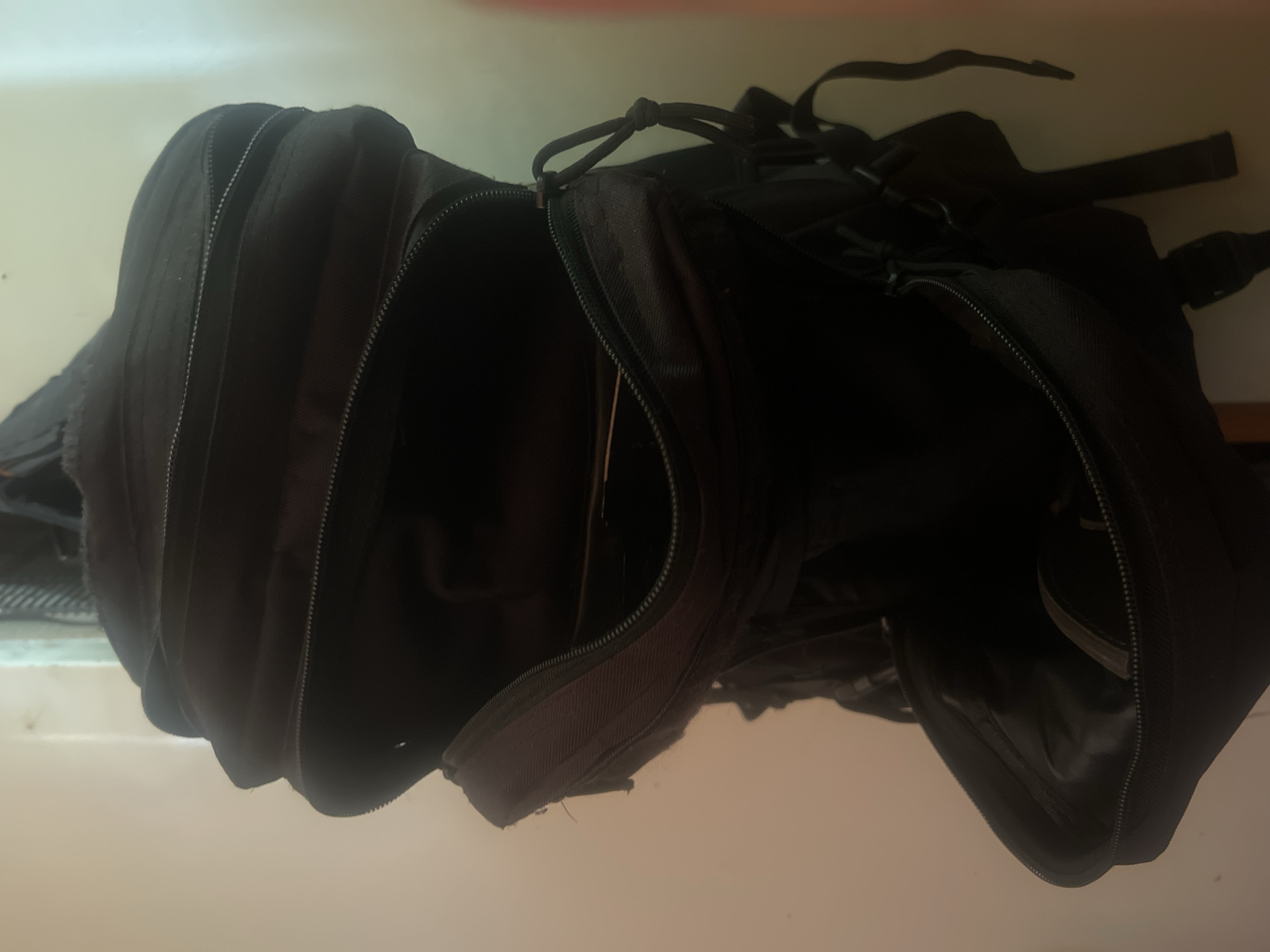 A black backpack is shown hanging against a wall, possibly in a room in Charentsavan, Armenia. The backpack is partially open, revealing its interior compartments. The lighting is dim, casting shadows and highlighting the textures of the fabric and zippers. There are no people visible in the image.