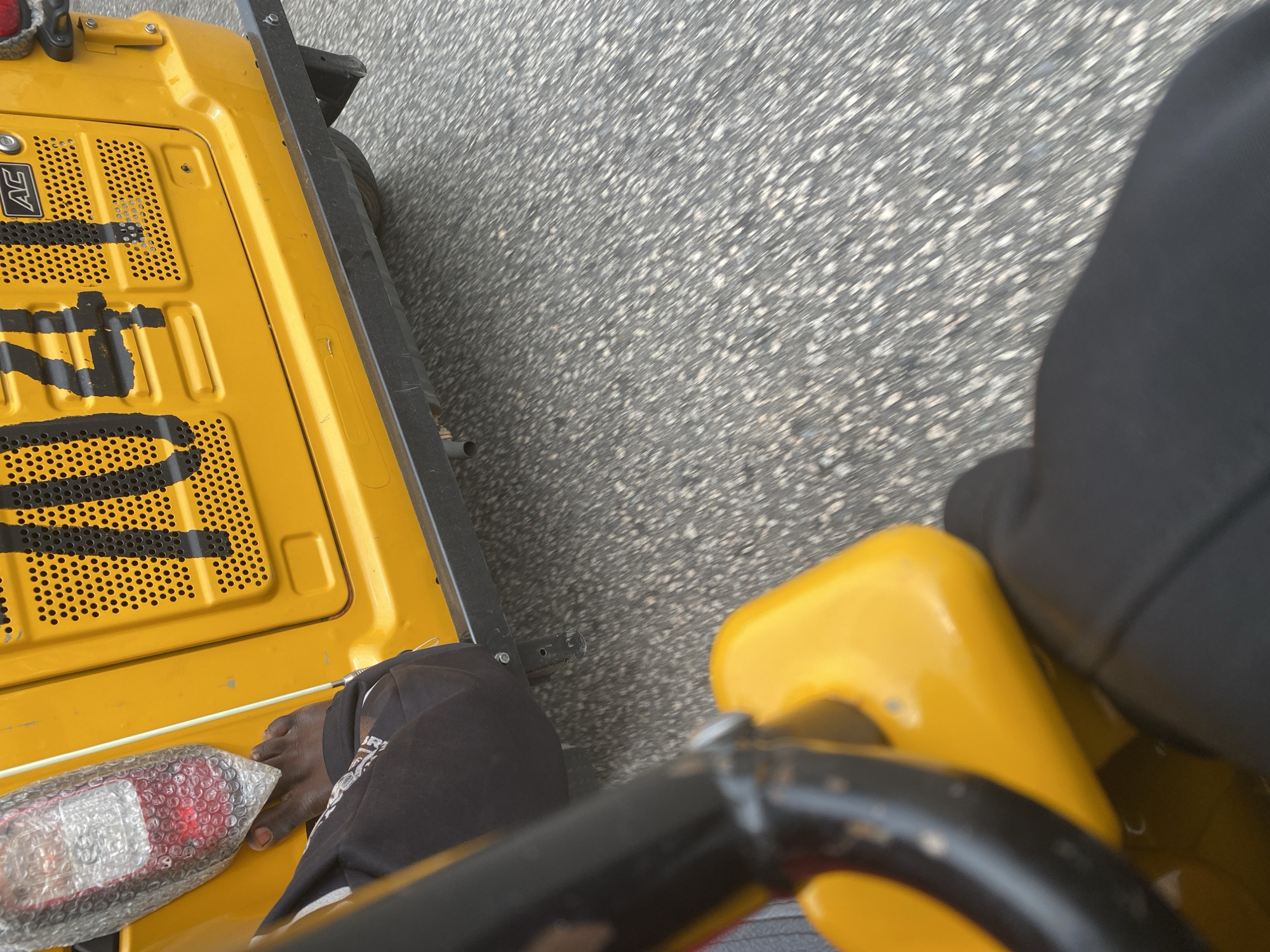 This media file shows a close-up, overhead view of a person's bare foot resting on the yellow surface of what appears to be a road roller or similar construction equipment. The foot, with visible toenails, is positioned near a red taillight encased in bubble wrap, which is also on the yellow surface.
The equipment is situated on a gray, asphalt-like surface, suggesting an outdoor setting such as a road or construction site. The scene is lit, indicating it is daytime. The background is blurred, but the texture of the ground suggests a slightly uneven surface.
The person's leg, clad in black fabric, is visible. The angle of the shot and the blurred background hint at movement, possibly the equipment being transported or operated. There is no discernible text in the visible portion of the image, except for what might be a faded logo or lettering on the yellow surface.
The overall impression is functional and utilitarian, focused on the equipment and the person's interaction with it in what is likely a work environment. Given the provided location context of Jalingo, Nigeria, the scene is situated in that geographical area.