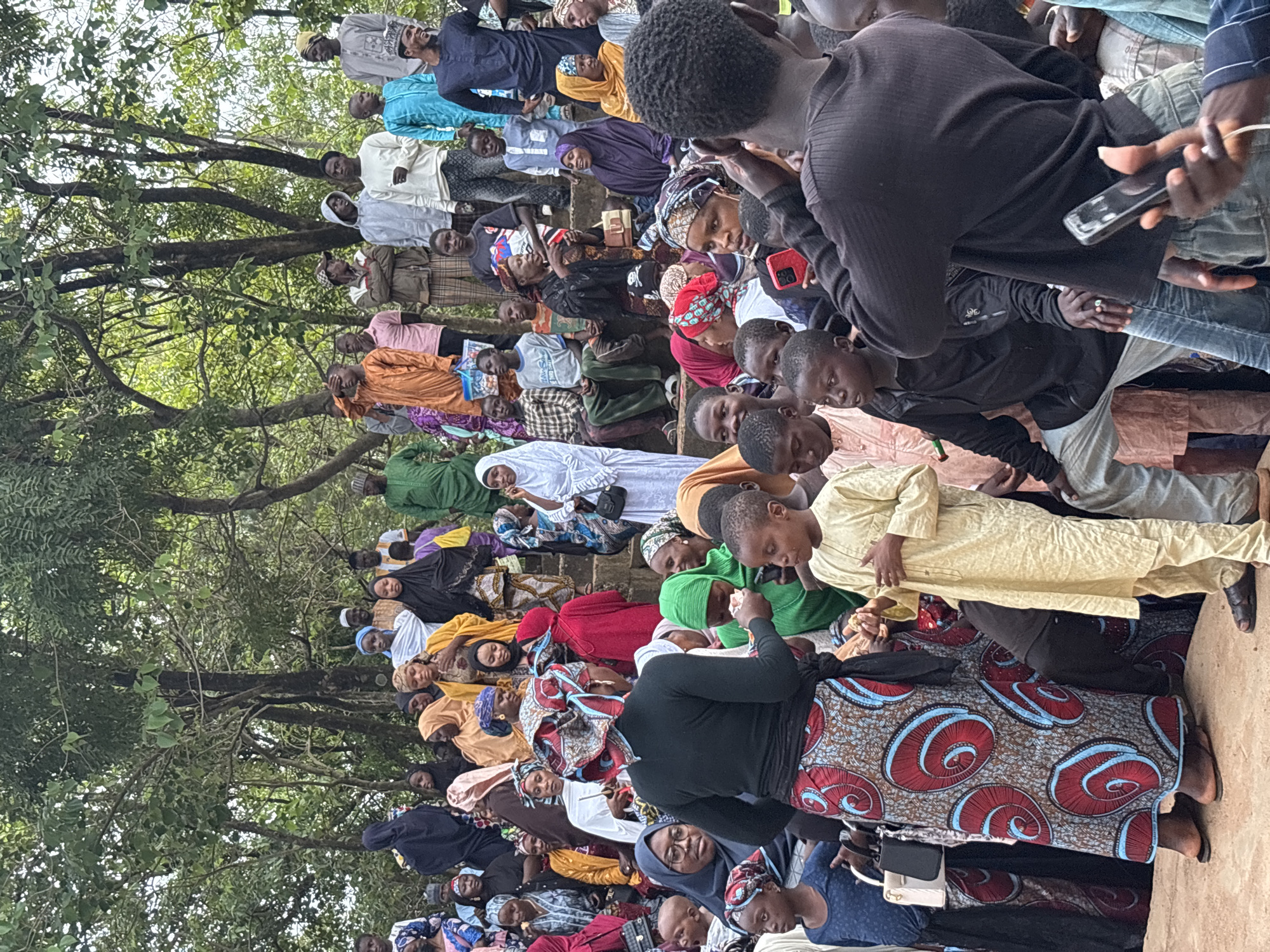 The image depicts a large outdoor gathering of people in a tiered, natural amphitheater setting, likely within the city of Jos, Nigeria. A dense crowd of men, women, and children are visible, many wearing traditional Islamic attire, including hijabs, abayas, and kaftans in a variety of colors and patterns. The environment is lush with green foliage and trees, suggesting a temperate climate.
In the foreground, a woman in a black top and a vibrantly patterned skirt with large red and blue spiral designs is facing away from the camera. She appears to be interacting with a young boy in a light yellow traditional outfit, who is holding something in his hands. Behind them, other individuals are looking in various directions, with some engaged in conversation.
Further back, the crowd is densely packed on stepped seating that appears to be carved into the natural landscape. The people in this area are a mix of ages and genders. Many are looking towards a central point, possibly an event or a speaker not fully visible in the frame. The overall atmosphere suggests a community event, possibly religious or cultural. The lighting is diffused, indicating an overcast day or shade provided by the trees.