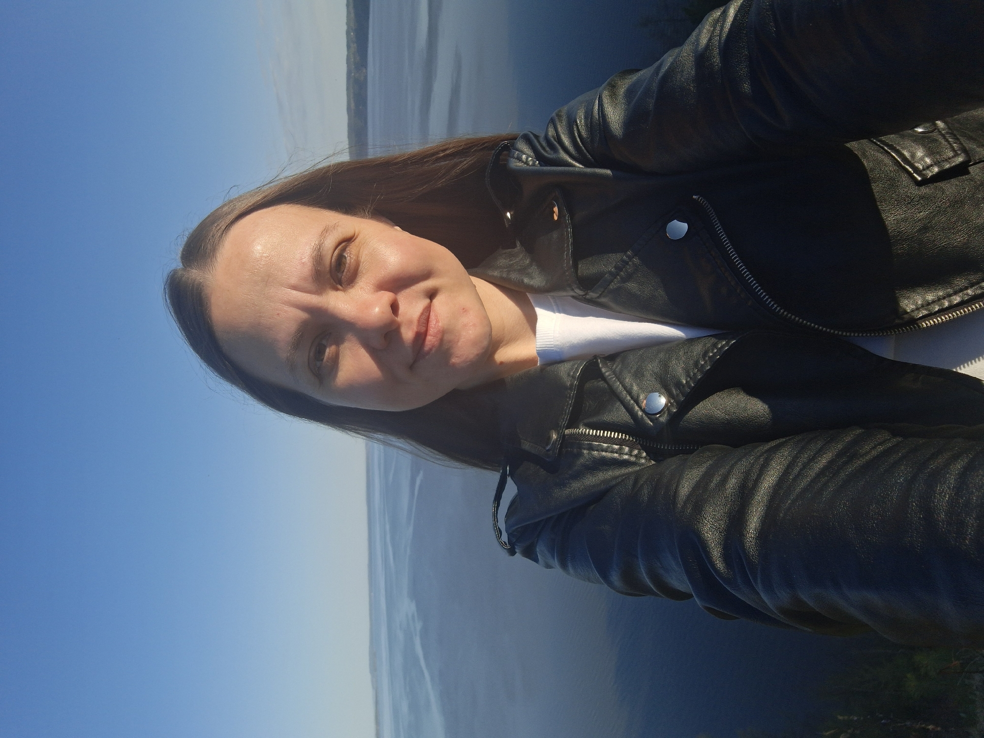 A woman is standing outside in Костоваты, Russia, taking a selfie. She has brown hair and is wearing a black leather jacket over a white shirt. In the background is a body of water that appears to be a lake or a river. The sky is blue with some haze. The overall scene is outdoors in daylight.