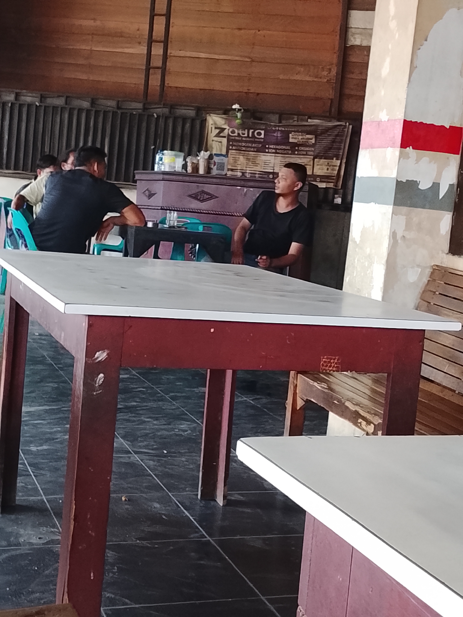 The image captures an indoor scene at a local eatery in Lhokseumawe, Indonesia. In the foreground, a white-topped table with a dark red base is prominent. In the midground, three individuals are seated at tables. Two men, one wearing a black t-shirt and the other a light-colored shirt, are facing away from the camera. A third man in a black t-shirt is seated facing towards the camera, looking slightly to his left. Behind them, a long, dark table is visible, along with several chairs, some in bright green. A banner for "Zaura" is mounted on the wall behind the seated individuals. The environment appears rustic, with wooden elements and a concrete floor. The overall atmosphere suggests a casual gathering or dining experience.