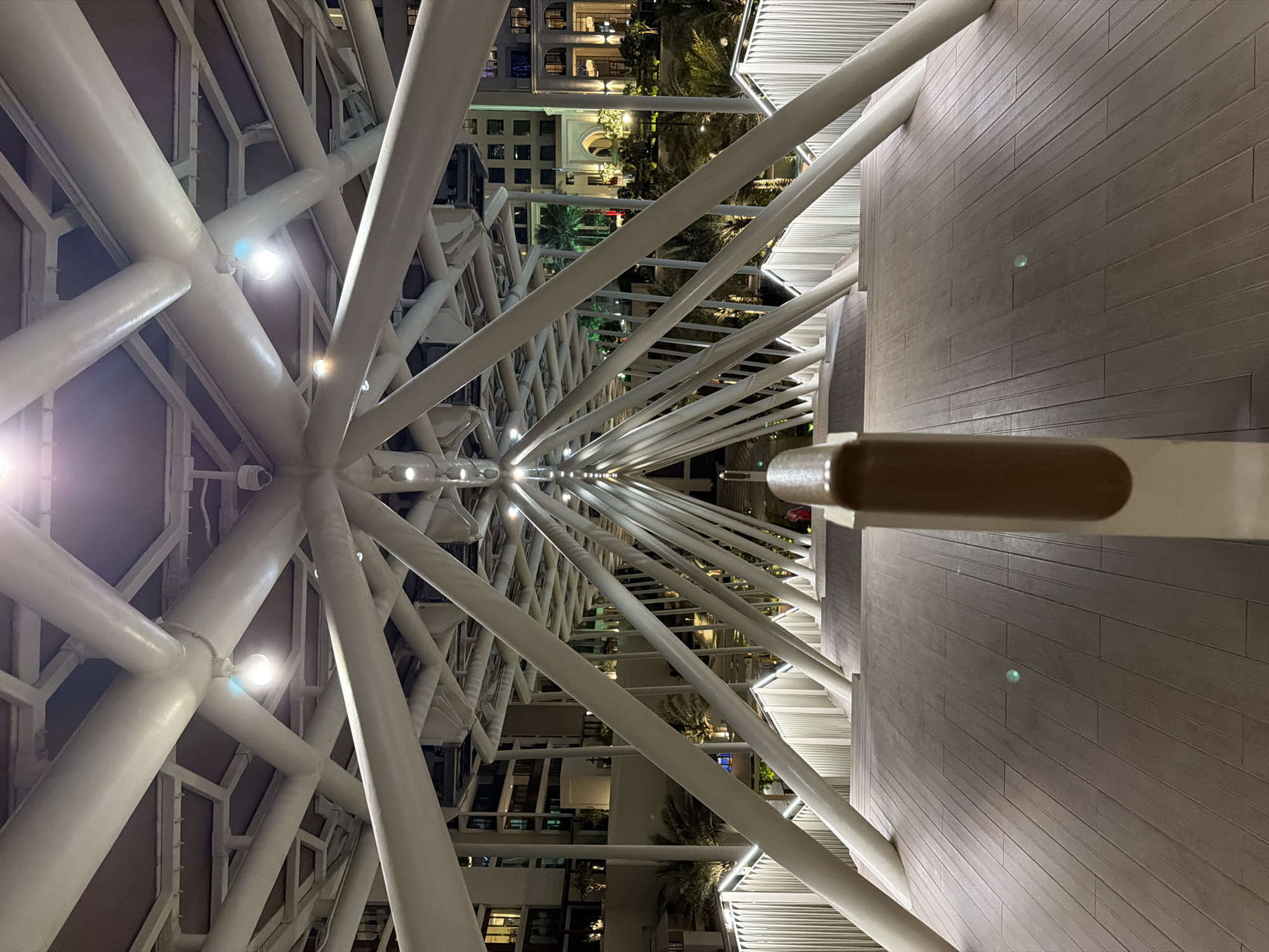 This nighttime image captures an upward perspective of a structure in Dubai, United Arab Emirates. The primary focus is on an intricate framework of white, cylindrical beams that converge towards a central point. Small lights are mounted at the joints of the structure. Between the beams, the cityscape is visible, including buildings with lit windows and lush palm trees. To the right of the frame, a wooden walkway with horizontal planks is partially visible. A section of a white railing with a tan-colored handrail is in the foreground, partially obstructing the view of the structure.