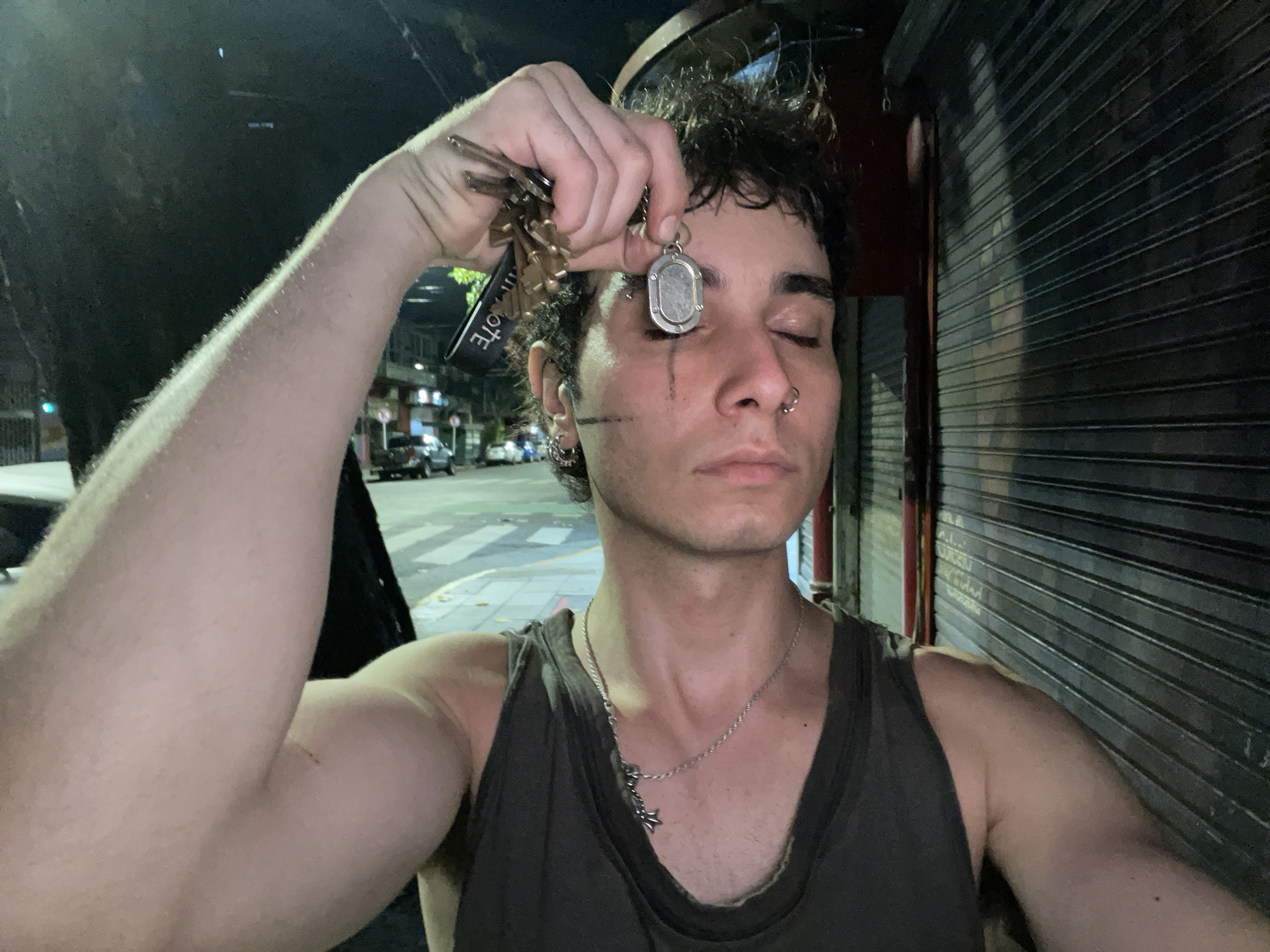 A person is standing on a sidewalk at night in Buenos Aires, Argentina. The person is fair-skinned, with dark, curly hair and a thin, athletic build. They have a nose ring in their left nostril and are wearing a silver chain necklace. They have a straight, dark, vertical line drawn on their left cheek extending from the corner of their eye down to the cheek. The person's eyes are closed, and they are holding a silver pendant on a chain in front of their left eye with their right hand. They are also holding a set of keys on a chain with a black fob in their right hand. They are wearing a sleeveless dark grey or brown shirt. To their right, a metal roll-up door is in the background. Across the street, there are cars parked along the sidewalk, and a street sign with the word "COSTE" is visible. The street appears to be wet from recent rain. The image is lit by streetlights, creating a slightly grainy, low-light effect.