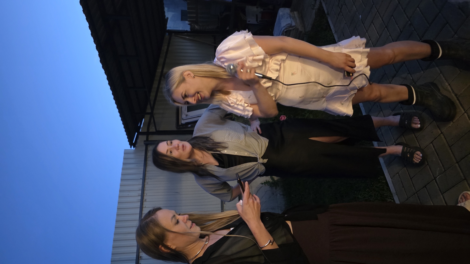 Three women are gathered outdoors in what appears to be a backyard or patio area in Bulgakovo, Russia, during twilight. The sky is a deep blue, suggesting dusk.
On the left, a woman with blonde hair is looking down at her smartphone, which she is holding in her right hand. She is wearing a black shirt with a silver necklace and a long, dark skirt.
In the center, a woman with dark brown hair is wearing a striped, open-collared shirt tied at the waist, over a black slip dress. She has a black skirt with a high slit. Her hands are on her hips, and she is looking towards the woman on the right.
On the right, a blonde woman is holding a microphone in her right hand and appears to be speaking or singing, as she is looking towards the other two women and smiling broadly. She is wearing a pale pink dress with ruffled sleeves and a ruffled hem. She is also wearing black rubber boots.
The background includes a light-colored corrugated metal fence to the left, and a dark-colored roofed structure to the right, possibly a carport or gazebo. The ground is paved with interlocking grey bricks. Some greenery is visible in the background.