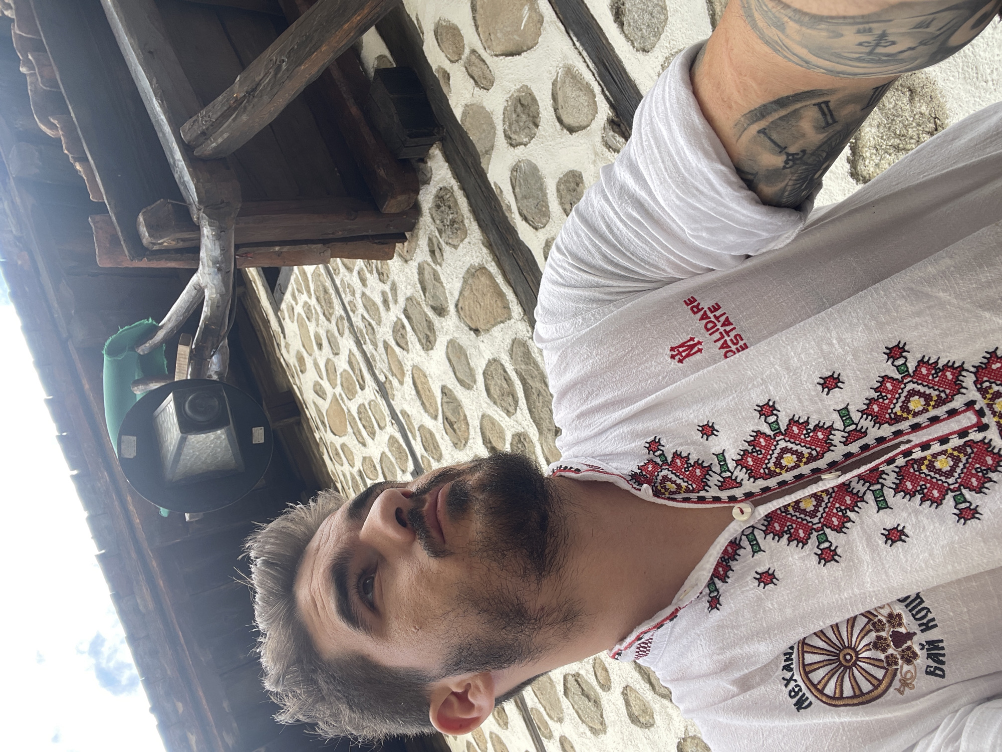 A male individual is depicted from a low-angle perspective, looking upwards and to the right, against a stone and timber building facade. He has a full dark beard, mustache, and short dark hair with visible grey strands. He wears a white, long-sleeved shirt with intricate red, white, and green traditional embroidery around the collar and chest opening. The shirt features an embroidered logo on the left chest (viewer's right) depicting a wagon wheel with Cyrillic text "МЕХАНА БАЙ КОЦЬ", and another logo on the right chest (viewer's left) displaying "DALIDARE ESTATE" with a stylized "M" above it. His left arm (viewer's right), with the sleeve rolled up, reveals a large, dark-toned tattoo that includes a segment of a clock face with Roman numeral II visible.
The background consists of an exterior building wall constructed from irregularly shaped, light-colored stones set in white mortar, horizontally intersected by dark wooden structural beams. Above the individual, a dark-colored, cylindrical light fixture or camera is affixed to a carved wooden beam extending from the structure. A piece of green fabric is draped over this wooden beam. Overhead, a section of a reddish-brown tiled roof is visible, with a small portion of an overcast sky in the upper left corner.