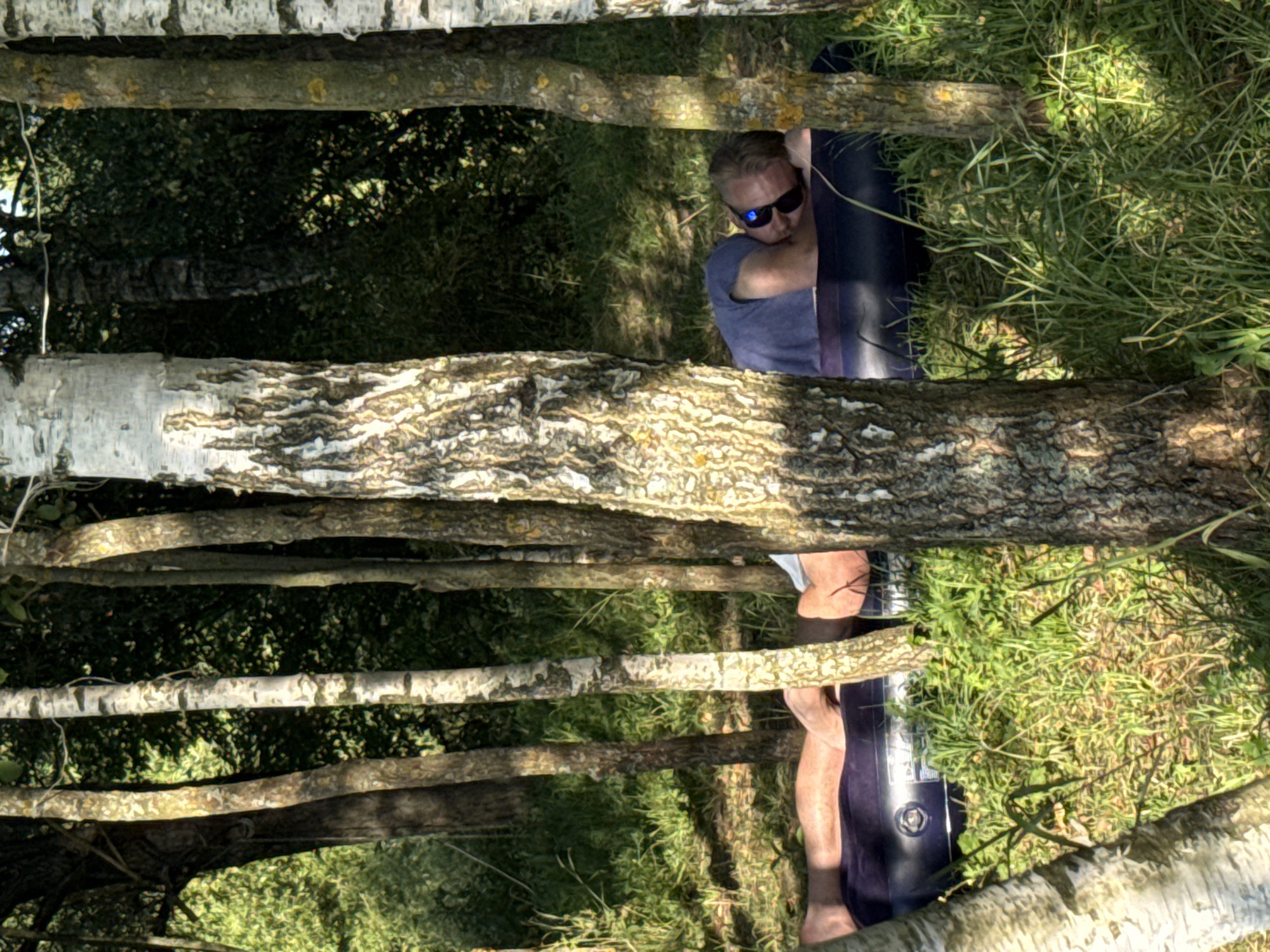 In the city of Kuzemkino, Russia, a man is seen lying on an inflatable mattress in a wooded area, resting on his side. He is wearing a blue t-shirt and dark sunglasses. The mattress is dark blue and appears to be partially deflated. The surrounding environment is characterized by a dense growth of trees with visible trunks and foliage, and lush green grass. Sunlight filters through the trees, creating patches of light and shadow on the ground. The image captures a moment of relaxation in a natural outdoor setting.