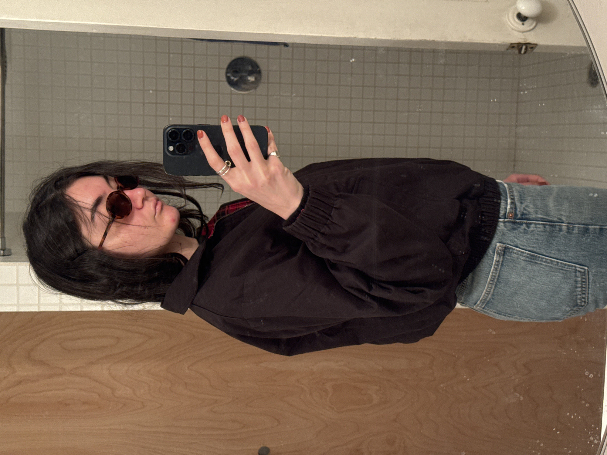 A person is captured in a mirror selfie, holding a black smartphone with their right hand to take the picture. The image is rotated 90 degrees clockwise, showing the person lying horizontally in the frame.
The individual has dark hair, styled down, and is wearing round, tortoise-shell sunglasses that obscure most of their eyes. Their lips are slightly pursed. They are dressed in a black bomber-style jacket over what appears to be a red and black plaid shirt, visible at the collar. The jacket has an elasticated hem and cuffs. Below the jacket, light-wash blue denim jeans are visible, featuring a back pocket. Several silver rings are worn on the fingers of the hand holding the phone, and their fingernails are painted an earthy orange or terracotta color.
The background, seen through the mirror's reflection, consists of small, light-colored square tiles, suggesting a bathroom or tiled wall. A circular drain or fixture is visible in the tiled wall. Above the tiled area, a white border or trim is present. In the upper right corner of the reflection, a white doorknob and a metallic hinge are visible, indicating a door within the reflected space. The bottom portion of the image shows a wooden surface, likely a sink counter or vanity. The overall lighting suggests an indoor setting, possibly with artificial light. No visible text is present in the image. The location is contextualized as New York, United States of America.