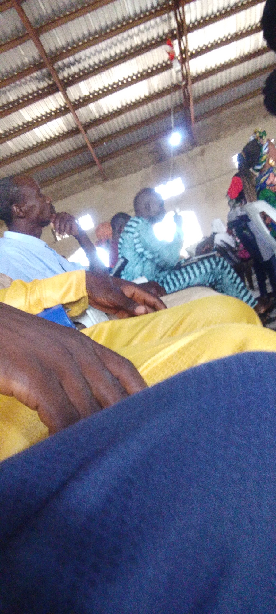 The image depicts an indoor scene, likely a gathering or meeting, with a corrugated metal roof visible overhead. Several individuals are present, some in the foreground and others further back. In the foreground, a person's hands are visible, resting on what appears to be a yellow garment and a blue fabric. To the left, a man in a light blue shirt is seen in profile, with his hand near his face, suggesting he might be listening or in thought. Further back, another individual is wearing a blue and black striped garment. Other people are partially visible in the background. The lighting suggests artificial light sources within the space. The scene appears casual and communal.