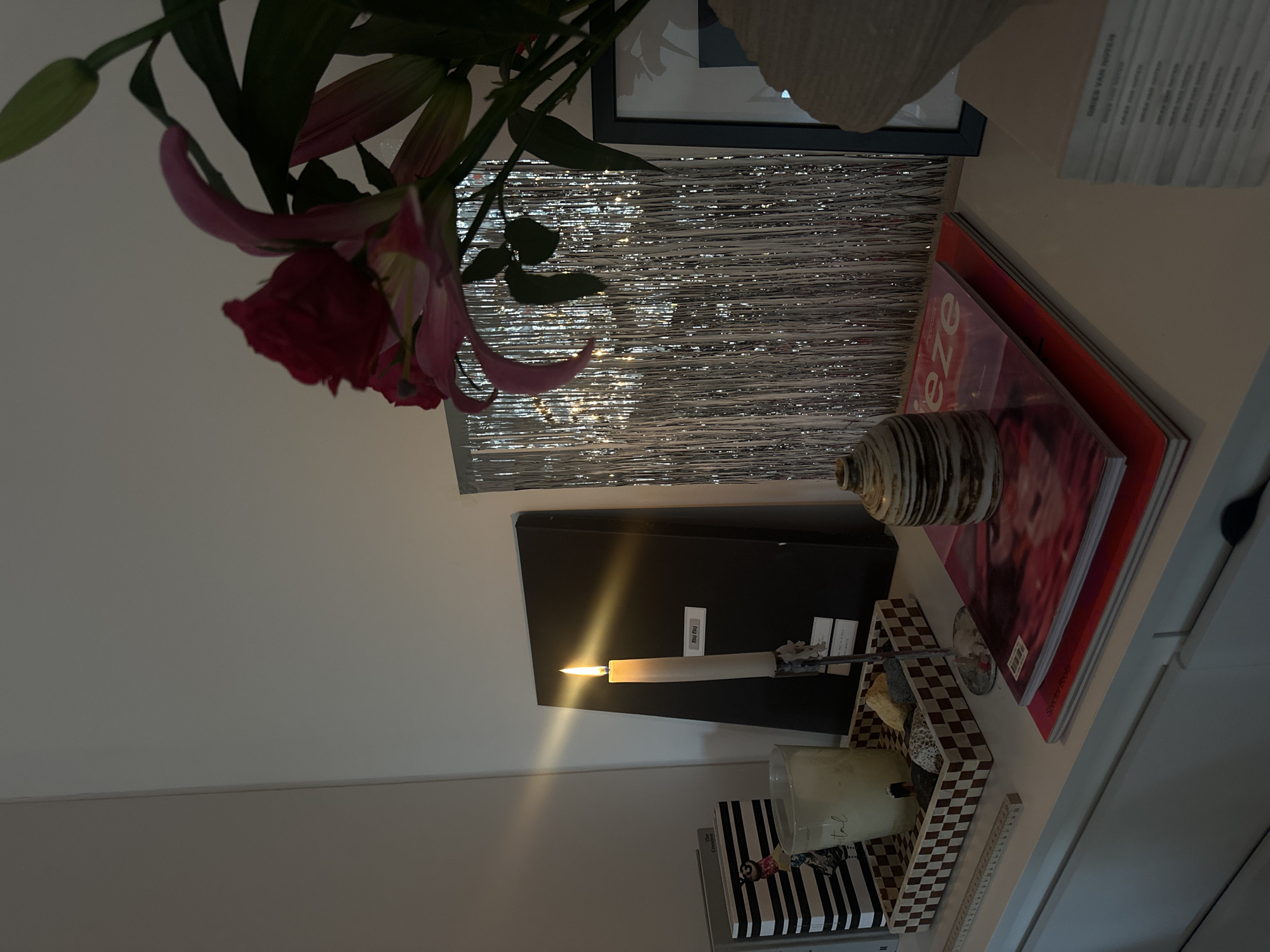 The image depicts a still life arrangement on a shelf, likely indoors. Prominent features include: a bouquet of flowers with red and pink petals and green foliage; a frame with a silver, tinsel-like backing; stacked books or magazines, one with the word "EZE" visible; a decorative ceramic vase; a lit candle in a metal holder positioned in front of a black box with a "Miu Miu" label, a patterned tray with stones and a clear glass jar candle; striped book. The overall environment suggests a personal space, possibly in a home in London, United Kingdom. However, the image doesn't offer explicit identifiers to confirm the location. No people or interactions are directly visible.