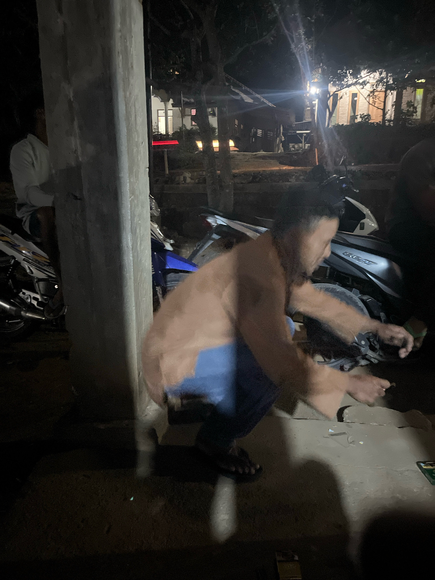 At night, in an outdoor setting in Kebumen, Indonesia, a shirtless man wearing blue pants is crouched down next to a motorcycle. His body is blurred due to movement. He appears to be interacting with something on the ground in front of the motorcycle, possibly with his hands. A portion of a concrete pillar is visible to the left of the frame, and other motorcycles and indistinct figures are present in the background, along with illuminated buildings and trees. The ground is paved and appears to be made of concrete or stone.