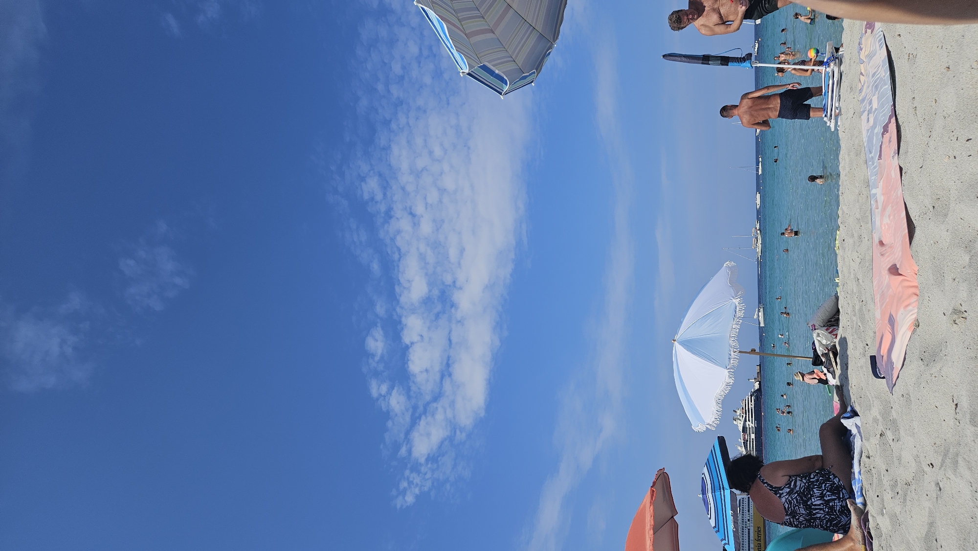 This image depicts a sunny day at a beach, likely in L'Île-Rousse, France, given the context provided. The scene is dominated by a clear blue sky with scattered white clouds. In the foreground, a sandy beach is populated with several beach umbrellas, including a large blue and white striped umbrella with fringe, and an orange umbrella. Beachgoers are visible in the water, some swimming and others wading. On the sand, a woman in a patterned swimsuit sits on a towel, facing away from the camera, under the shade of an umbrella. In the background, a large cruise ship is anchored in the sea, along with several sailboats. Two men are standing on the beach near the water, one of whom is shirtless. The water is a vibrant turquoise, and the overall atmosphere suggests a leisurely summer day.
